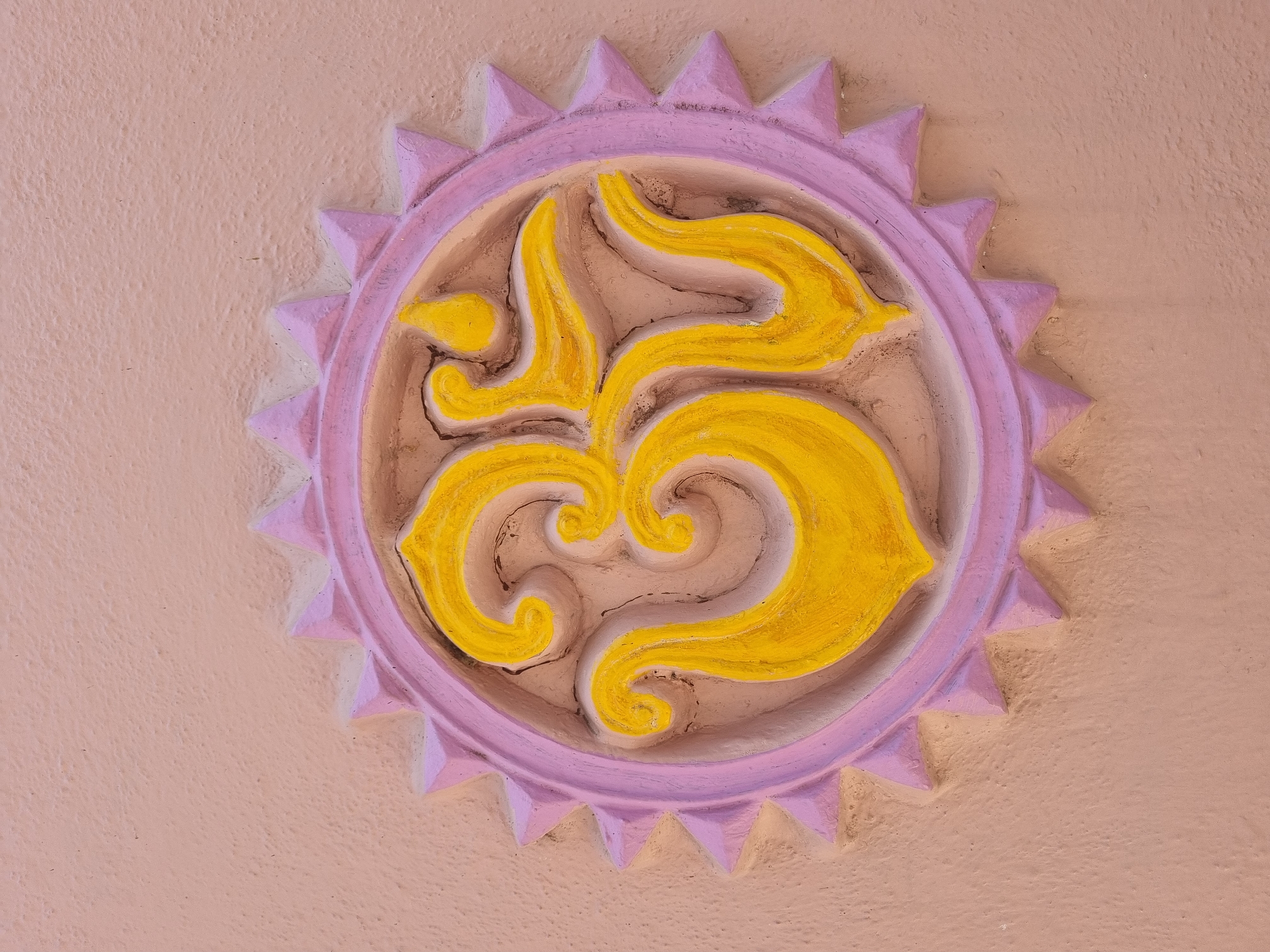 A relief sculpture depicting the Om (Aum) symbol is prominently displayed on a textured wall in Pedda Kammavaripalli, India. The Om symbol is rendered in bright yellow, deeply set within a recessed, light beige oval base. This oval is encircled by a lavender-purple frame featuring a jagged, sun-ray-like outer edge composed of triangular projections. The background wall surface is a uniform pale peach or light pink, exhibiting a textured finish. No individuals, actions, or other objects are present in the scene.