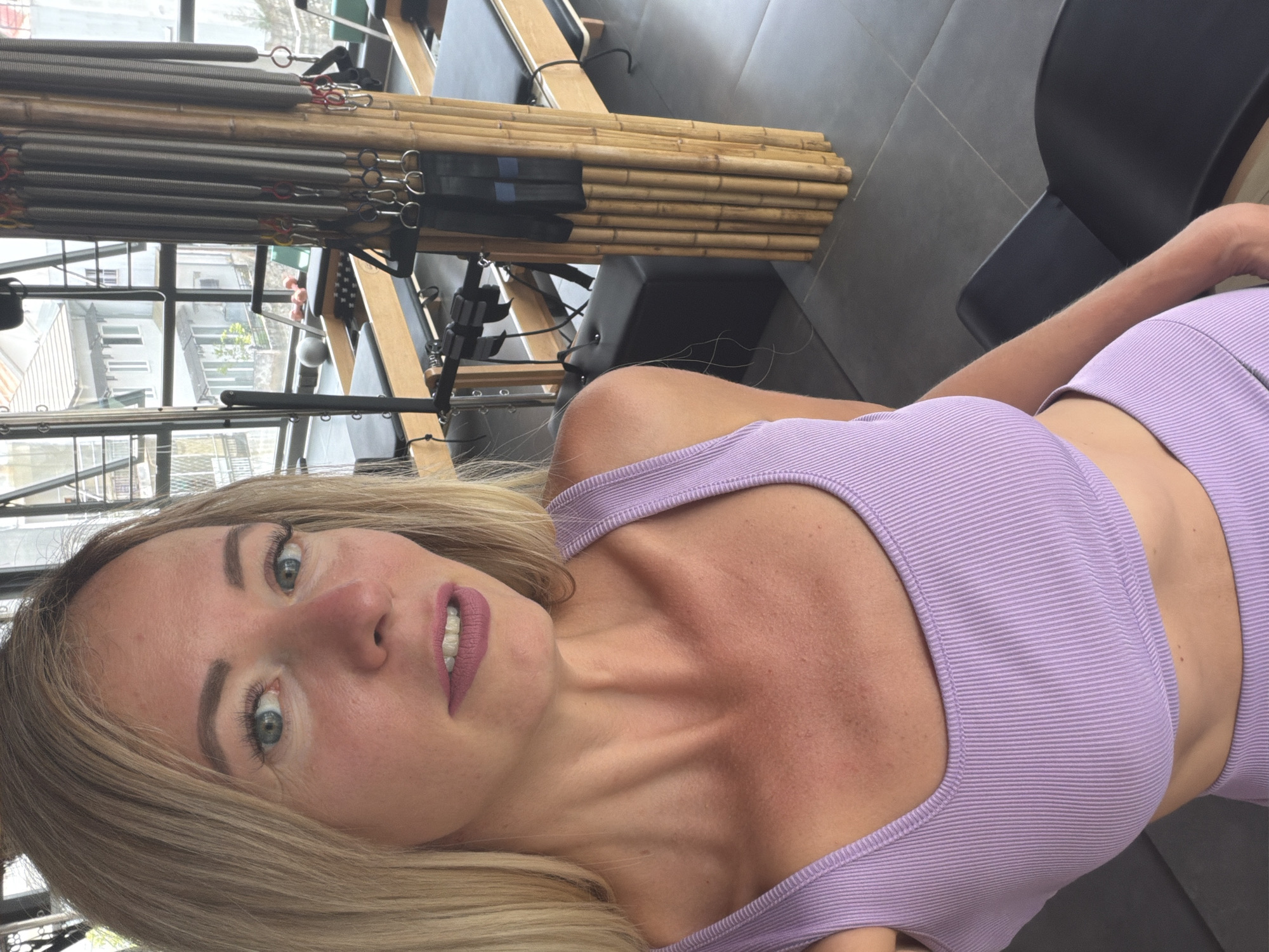 A woman with blonde hair and blue eyes, wearing a light purple athletic top and matching leggings, takes a selfie in a Pilates studio. The studio has large windows overlooking buildings and trees. Pilates equipment, including reformers and various attachments, are visible throughout the space. The floor is dark grey tile, and a bamboo-covered pillar stands near some of the equipment. The woman's facial expression is neutral. The lighting suggests it is daytime. The location of this studio is in Da Lat, Vietnam.