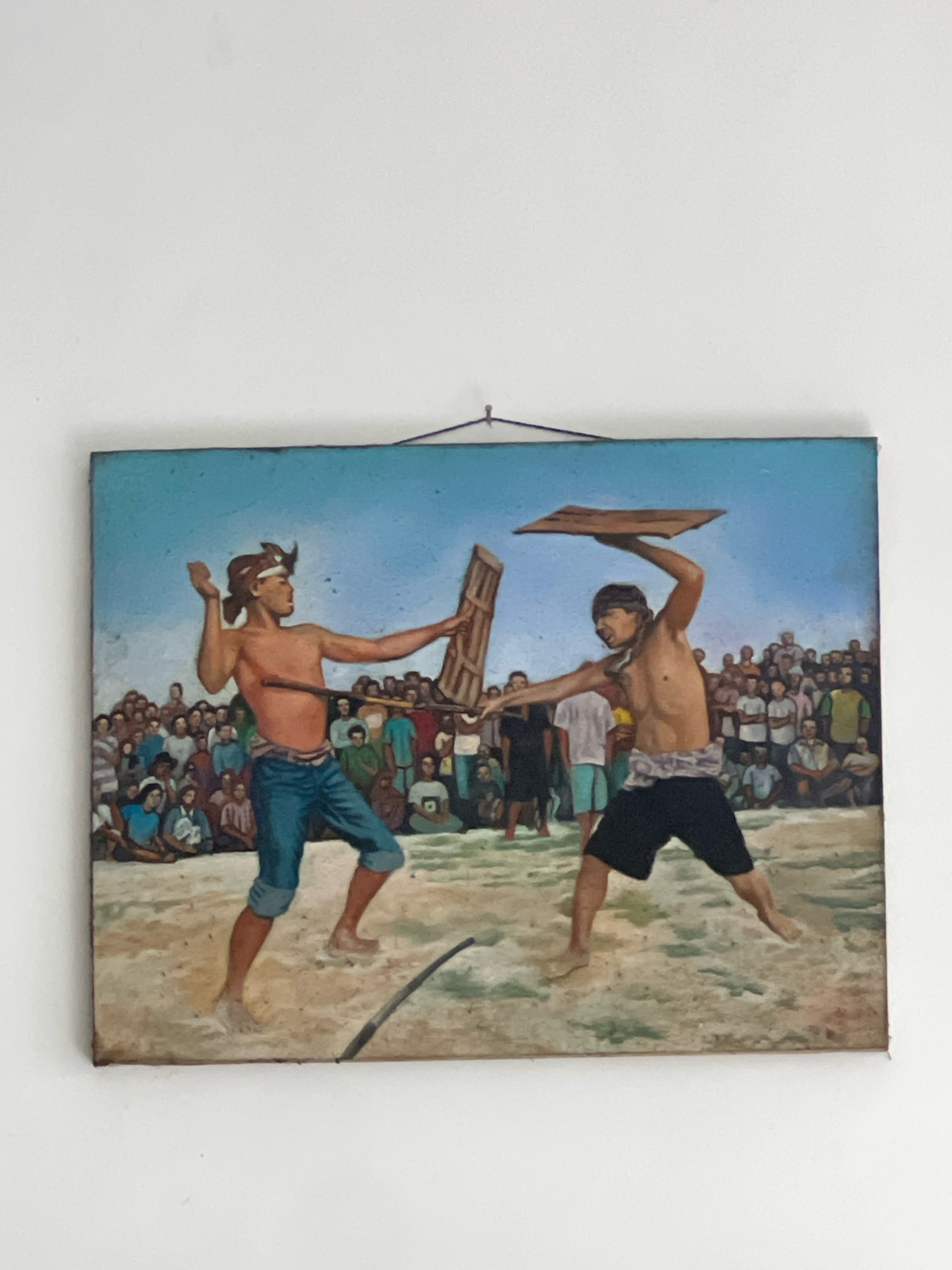 This is a painting depicting a scene of men engaged in a form of combat or ritual. Two men are prominently featured in the foreground, shirtless and wearing traditional lower garments. The man on the left is raising his right arm as if to throw an object, while holding a flat wooden shield in his left hand. The man on the right is in a defensive posture, holding a similar shield above his head. A dark, curved object, possibly a stick or a weapon, lies on the sand in front of them. Behind the fighters, a crowd of onlookers is gathered, suggesting a public event or competition. The background features a bright blue sky and a sandy or dusty ground. The overall impression is that of a historical or cultural reenactment taking place in Kuta, Indonesia, a location known for its beaches and cultural tourism.