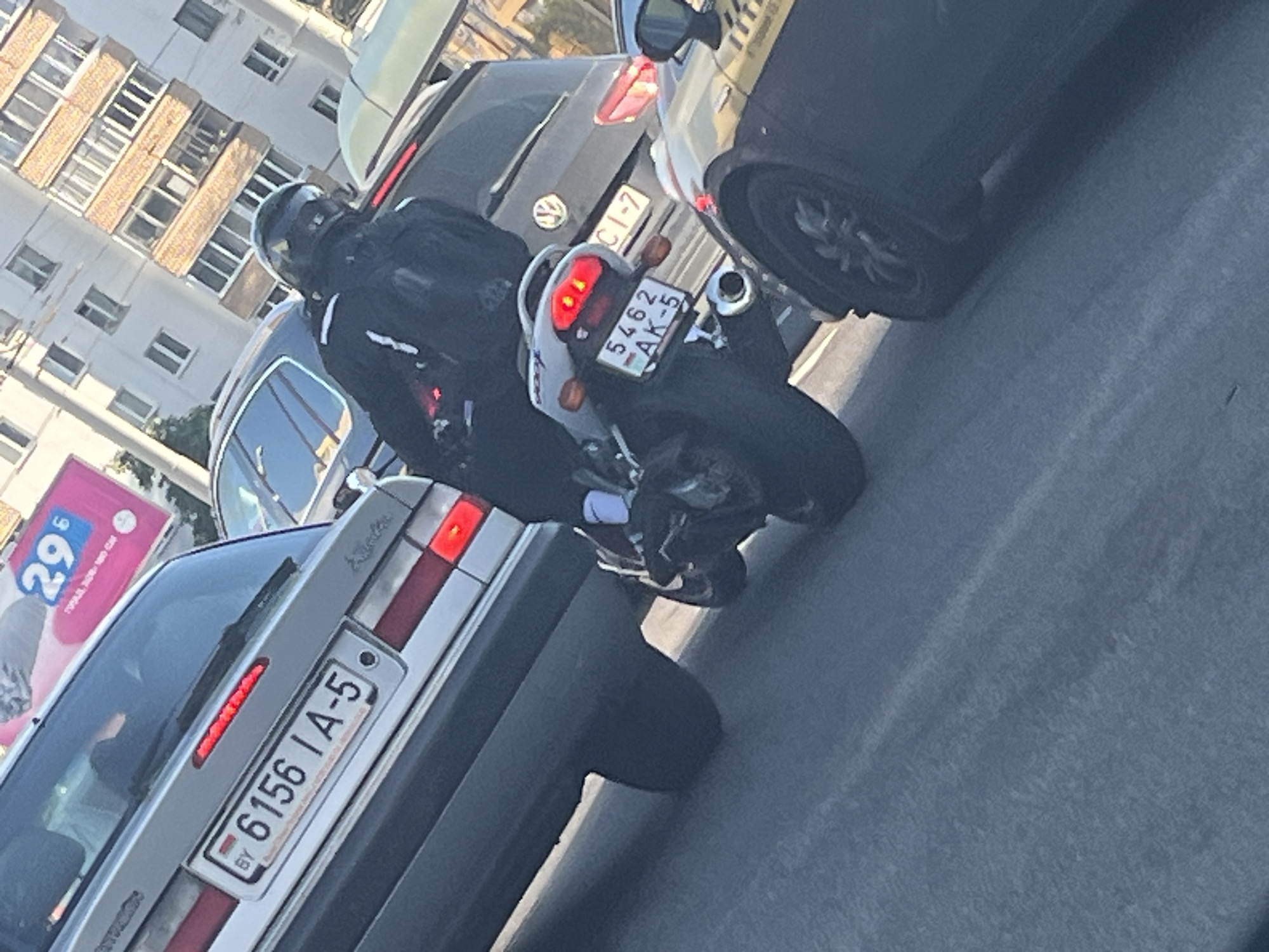 The image shows a traffic scene on a city street in Minsk, Belarus. A silver car with a license plate that reads "6156 IA-5" is in the foreground, with a portion of a billboard showing "29" and a pink design in the background. To the right of the car, a motorcycle with a rider wearing a black helmet and jacket is visible. The motorcycle's license plate reads "5462 AK-5". In the background, other vehicles and buildings are present, suggesting an urban environment. The lighting indicates it is daytime.