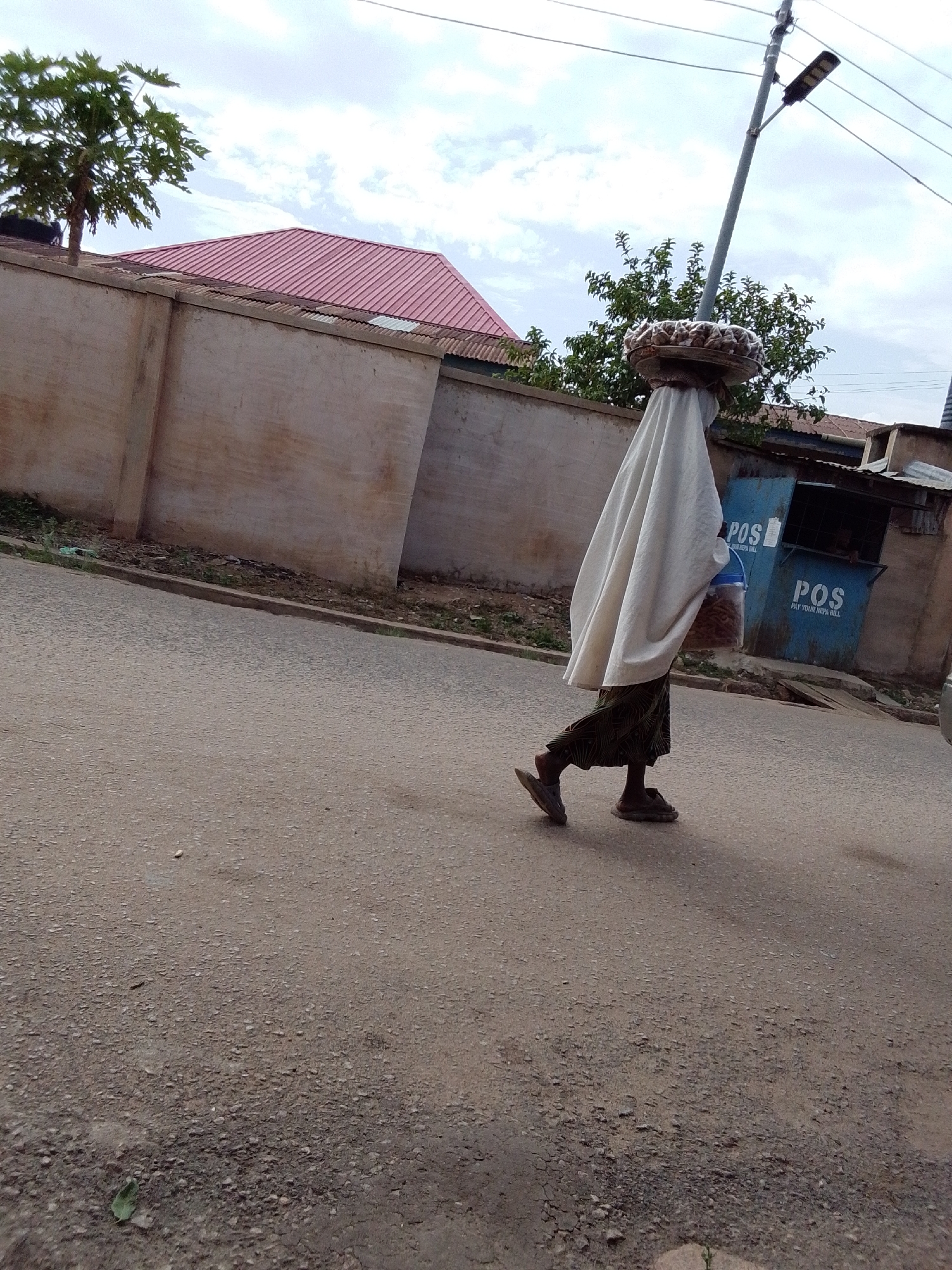 A person wearing a long white cloth walks along a paved street in an urban area of Unknown, Unknown. The person carries a tray on their head. The cloth covers their face and body. They wear sandals and a patterned skirt. A blue POS booth stands near the side of the street. A concrete wall and a house with a red roof are in the background. A utility pole rises near the person.
