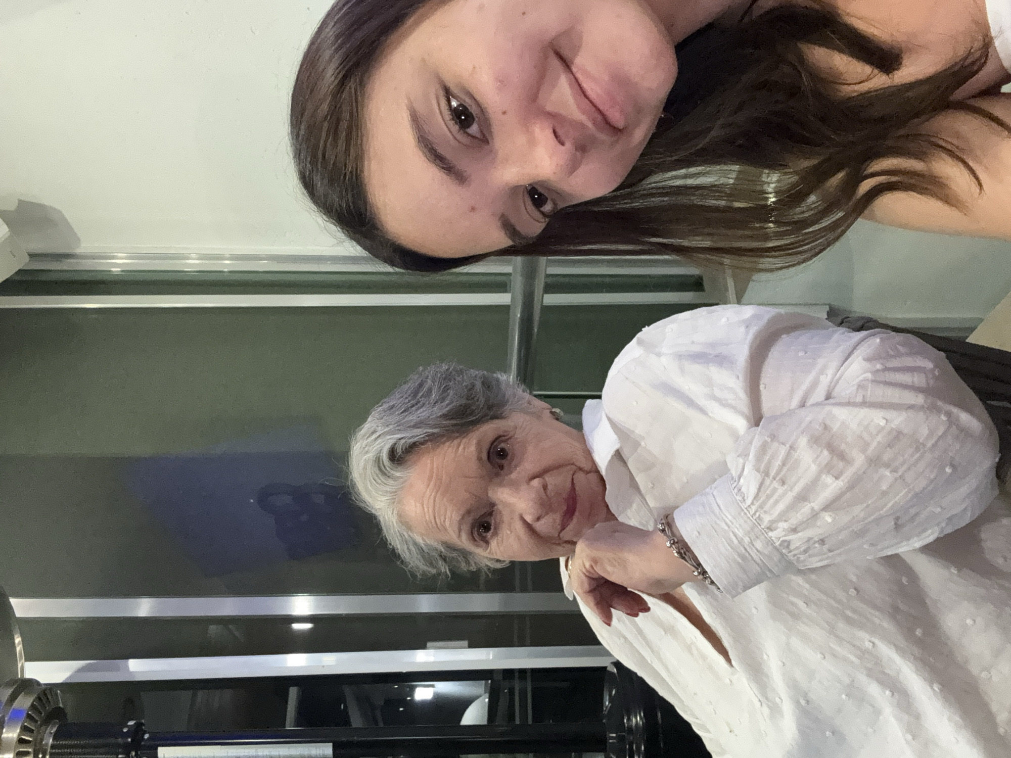 A selfie-style photograph, tilted slightly, features two women indoors. The younger woman is at the top, facing the camera with brown hair and fair skin. Below her is an older woman with gray hair and a white blouse. She is resting her chin on her hand and looking at the camera. In the background, there's a partially visible machine with metallic and black components, situated in front of a window or glass partition that reflects an unknown image. The location cannot be verified based solely on the image.