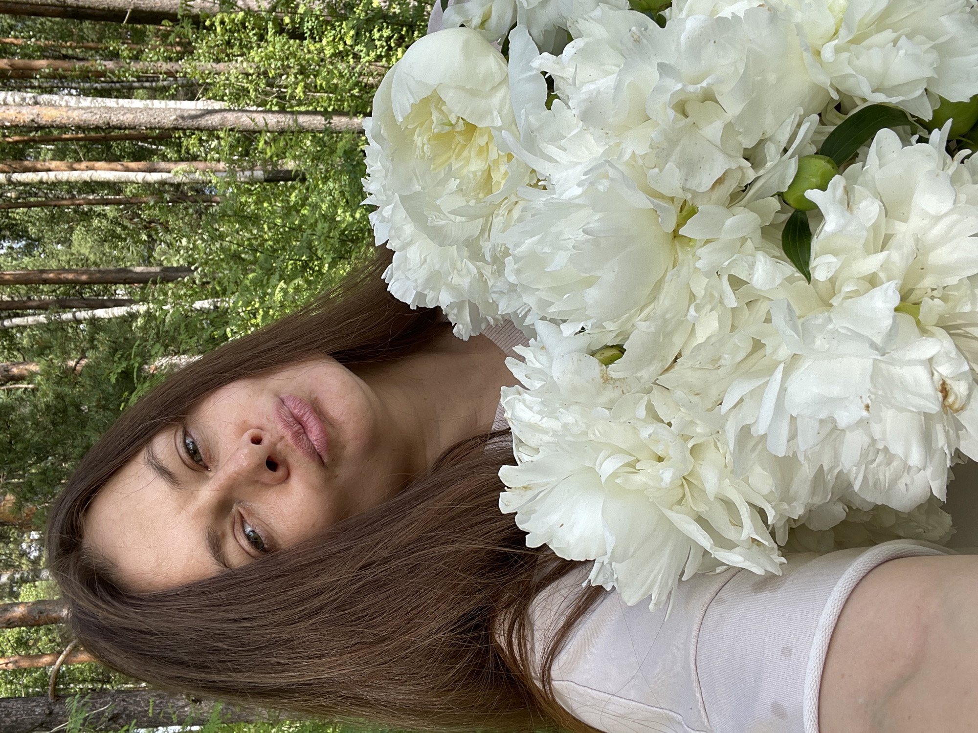 A woman with long brown hair is holding a bouquet of white peonies in front of her. She is wearing a light-colored t-shirt. The background consists of a forest with tall trees and green foliage, suggesting an outdoor setting. The lighting indicates it is daytime. The woman is looking at the camera with her lips puckered. The scene is captured from a medium close-up perspective. The location is not identifiable as Заречный, Russia, from the image itself.