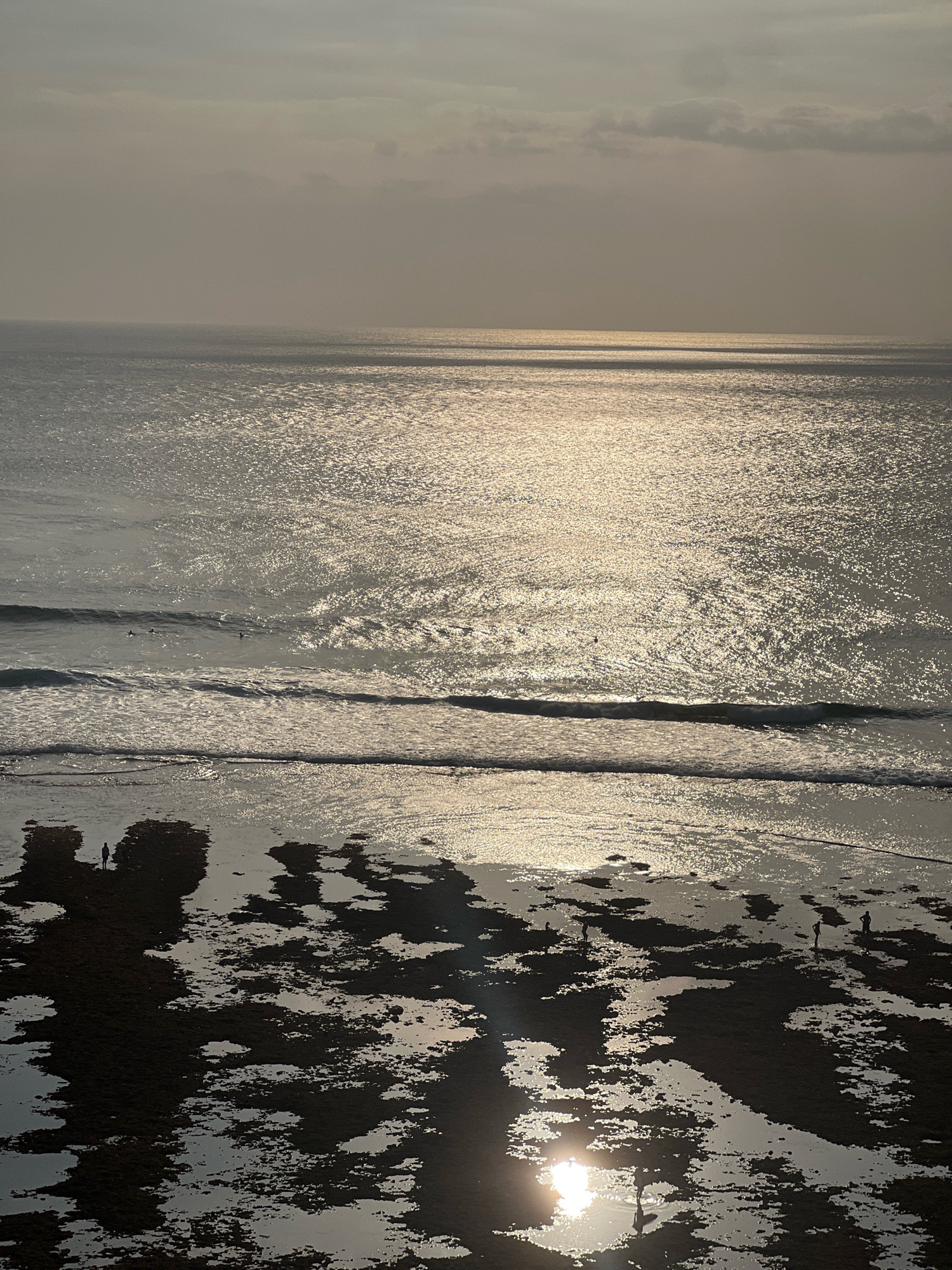The image captures a coastal scene in Kuta Selatan, Indonesia, likely during sunset or sunrise, given the low angle of the sun and its reflection on the water. The sky is overcast with diffused sunlight casting a shimmering, golden glow across the ocean's surface. Gentle waves are breaking near the shore, creating foamy white lines. The foreground depicts a rocky and tidal area, with dark, wet rocks interspersed with shallow pools of water that reflect the sky and sunlight. Several silhouetted figures are scattered across the beach and in the shallow water, appearing to be walking or standing. Further out in the ocean, a few surfers are visible on their boards, waiting for waves. The overall atmosphere is serene and tranquil, with the natural elements of water, light, and land dominating the composition.