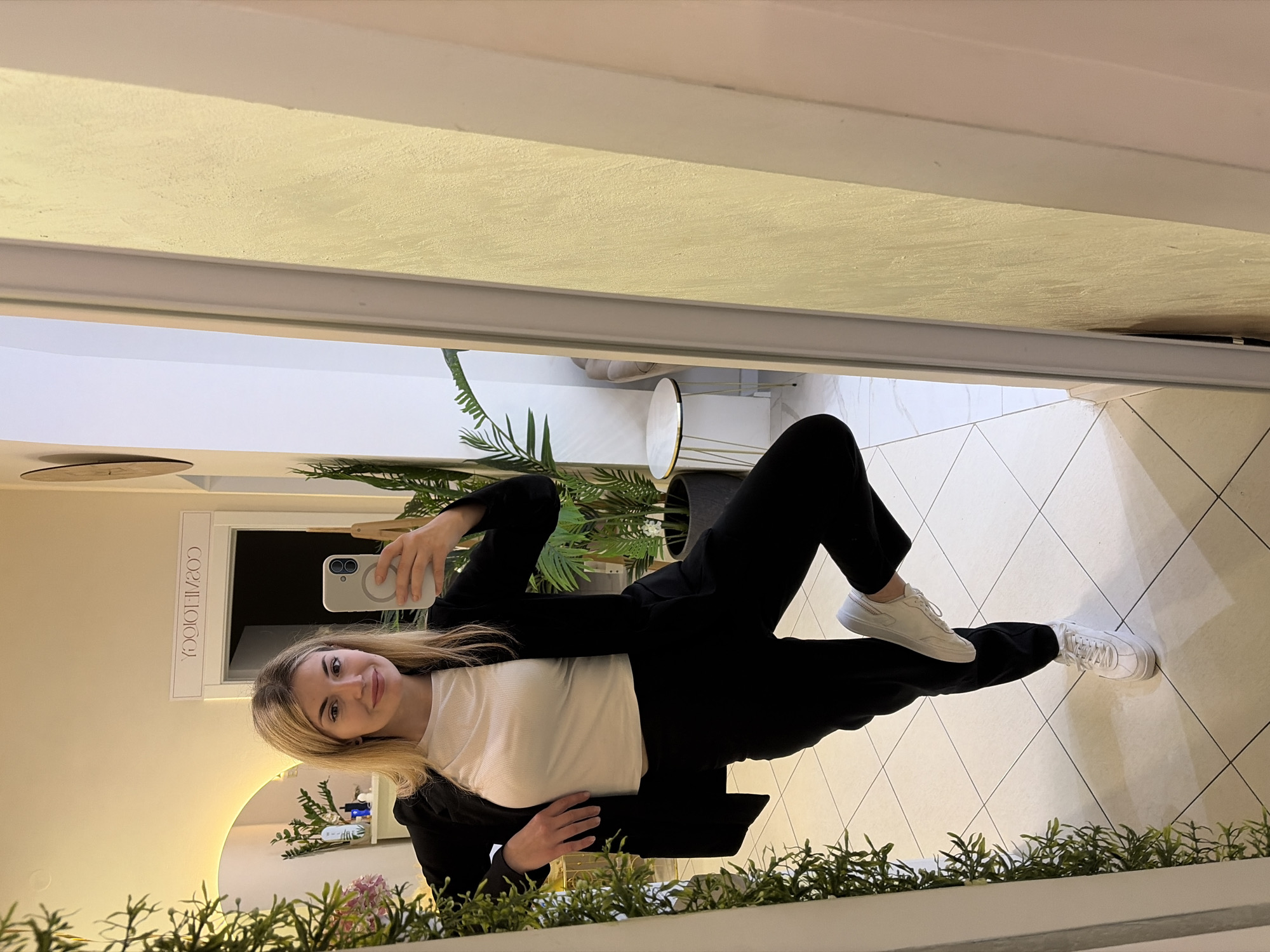 A young woman with blonde hair, wearing a white top and black blazer and trousers, is taking a selfie in a mirror. The mirror reflects a room with light-colored walls, a patterned floor, and greenery in the foreground. Reflected in the mirror is also the word "COSMETOLOGY" written backward. Outside the mirror, the ceiling is visible. It is likely the woman is in a business location. The photograph was taken in Budva, Montenegro.