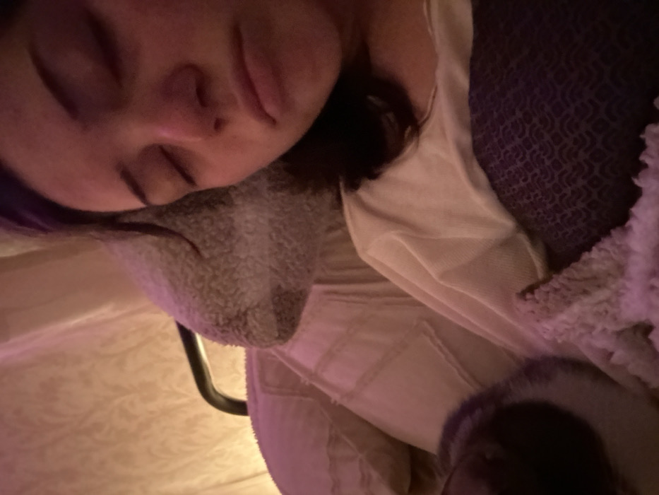 A close-up, low-angle shot depicts a person sleeping. The individual lies on their side, face visible, with eyes closed. They rest on a gray, plush pillow. Clothing and blankets in shades of white, beige, and a textured purple appear. A black metal object is also visible near the pillow. The lighting is soft and warm, casting a gentle glow on the scene.