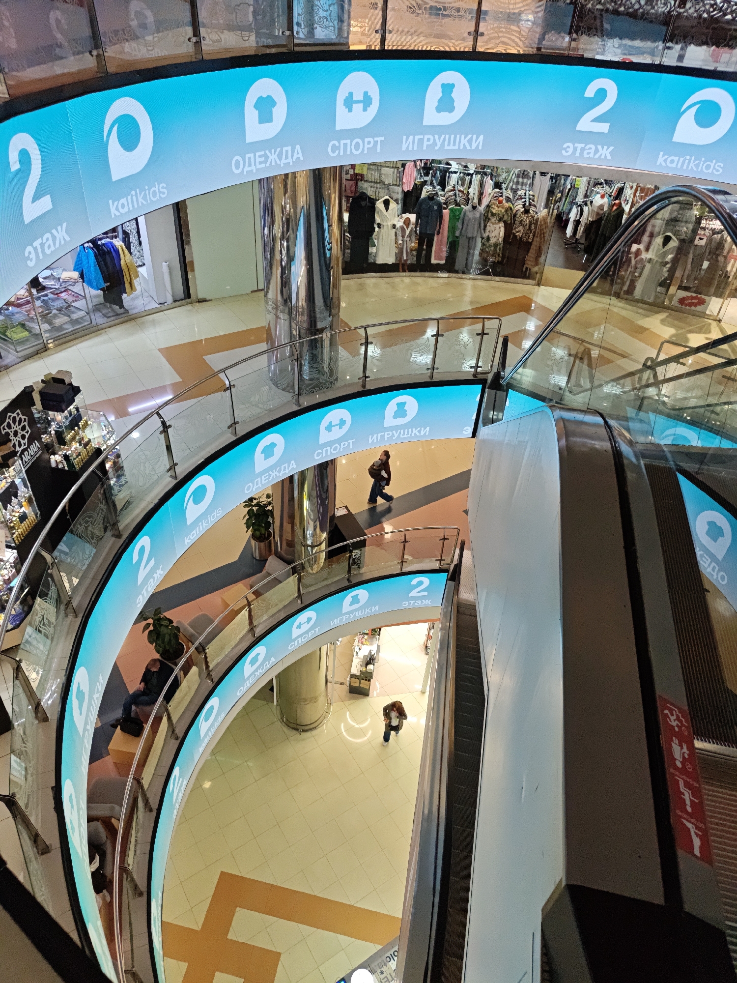 The image shows an aerial view of a shopping mall in Yekaterinburg, Russia. The interior features multiple levels, connected by an escalator and glass railings. On the railings are signs advertising Kari Kids and shops that sell clothing, sports apparel, and toys. People are walking or sitting in the mall. The floor has a geometric pattern of beige and orange tiles. The lighting is bright and appears to be artificial, suggesting an indoor environment.