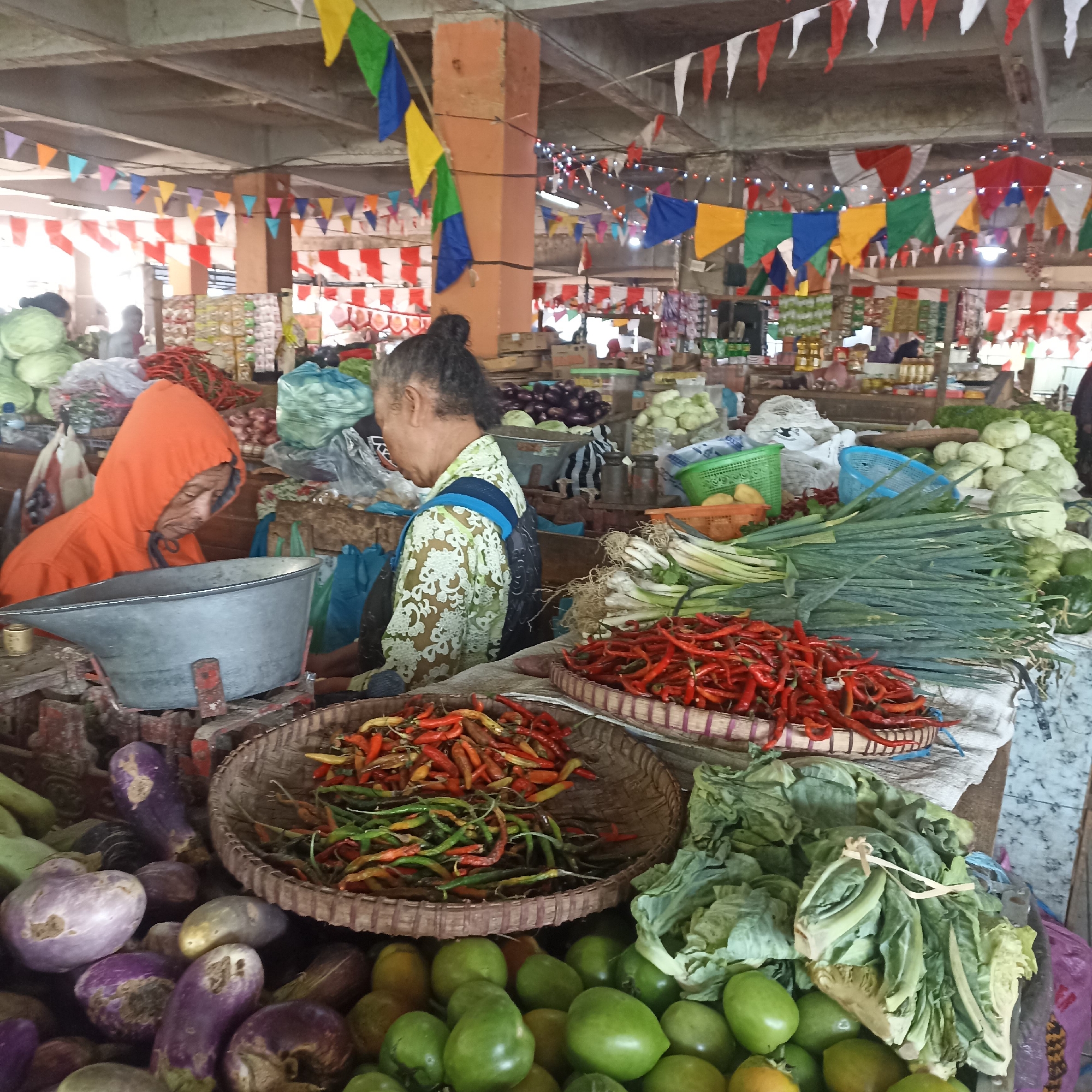 This image depicts an outdoor market stall in Magelang, Indonesia, decorated with colorful triangular flags. The stall is laden with a variety of fresh produce, including piles of red chilies in woven baskets, green onions, large cabbages, eggplants, and green tomatoes. Two individuals are visible; one, wearing an orange hooded jacket, is looking down at a large metal scale. The other, wearing a patterned green shirt, is also attending to the produce. The market appears busy, with other stalls and shoppers suggested in the background. The overall atmosphere is one of a vibrant, local marketplace.