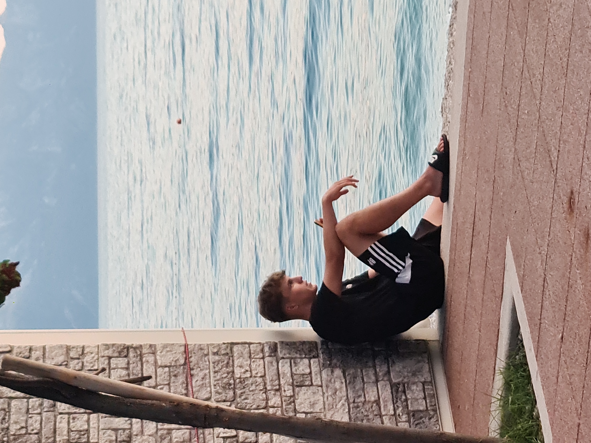 A young man with blonde hair sits leaning against a stone-tiled wall, facing right, with a body of water in the background. He is wearing a black t-shirt with white stripes on the sleeves and black shorts with white stripes on the sides. He is wearing black slides on his feet. He is holding a mobile phone in his right hand. The water appears calm with ripples, and a small orange buoy is visible in the distance. Mountains are faintly visible on the horizon. The ground in front of him is paved with reddish-brown tiles. A thin tree trunk is visible to the left of the man, in front of the wall. The scene appears to be an outdoor seating area near a body of water, possibly a promenade or waterfront in Općina Gradac, Croatia.