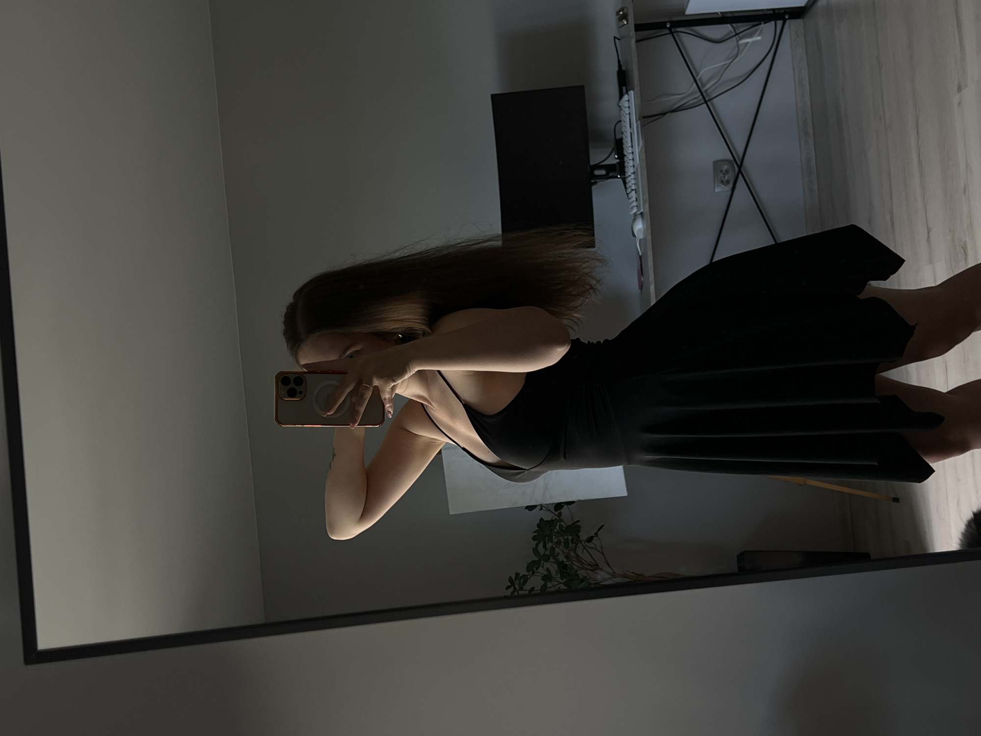 The image shows a woman taking a mirror selfie in what appears to be a home setting in Ternopil, Ukraine. She is wearing a black dress with spaghetti straps and a low neckline. Her long, brown hair is spread out. She is holding a smartphone with a rose gold case and a white circular logo, presumably a MagSafe accessory, in her left hand. A ring is visible on her left ring finger.
The background reflects a room with a wall on the left and a desk with a computer monitor and keyboard on the right. Wires are visible on the desk, and a power outlet is on the wall behind it. The floor is made of light-colored wood. A plant is visible in the reflection, suggesting an indoor environment. The lighting in the room appears to be artificial, as there are no clear indicators of natural light sources like windows or curtains. The overall mood is casual and private.