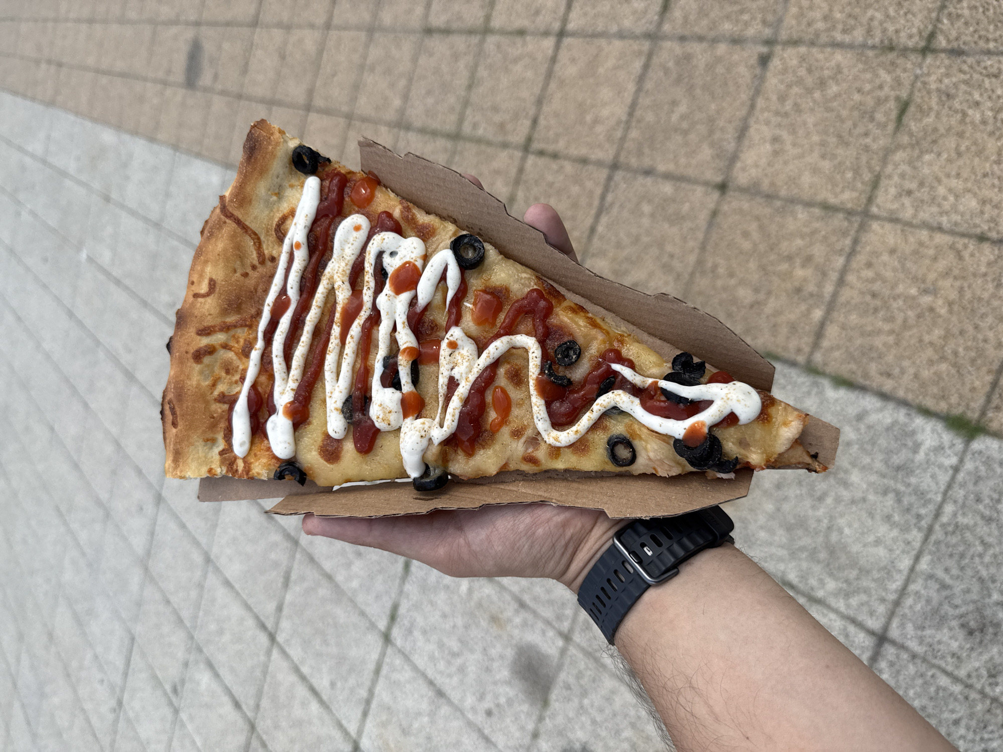 A slice of pizza is held in a person's hand against a backdrop of paved ground. The pizza is triangular, with a crust, cheese, tomato sauce, white sauce, olives, and what appear to be small diced red pieces. The person is wearing a black watch on their left wrist. The ground is composed of square paving stones. The image does not contain any information about the city of Vratsa, Bulgaria.