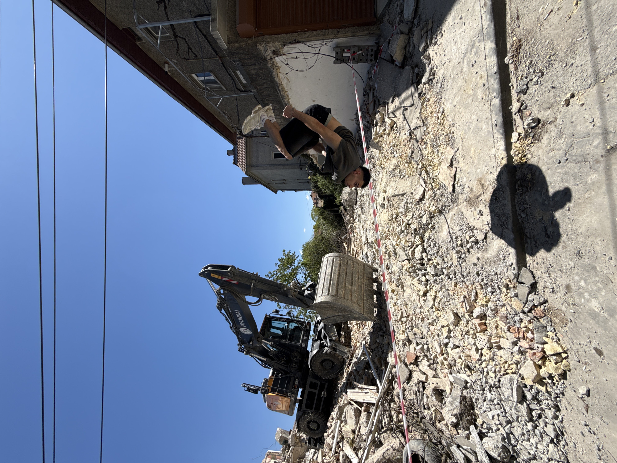 The image shows a scene of demolition in Shumen, Bulgaria. An excavator with the name "HYUNDAI" is visible on a pile of rubble. A man is doing a backflip near a building under demolition. There are trees and other buildings in the background. Overhead electrical wires are visible against a clear blue sky. The debris consists of bricks, concrete, and other construction materials. Red and white caution tape is visible. The photograph appears to be rotated clockwise 90 degrees.