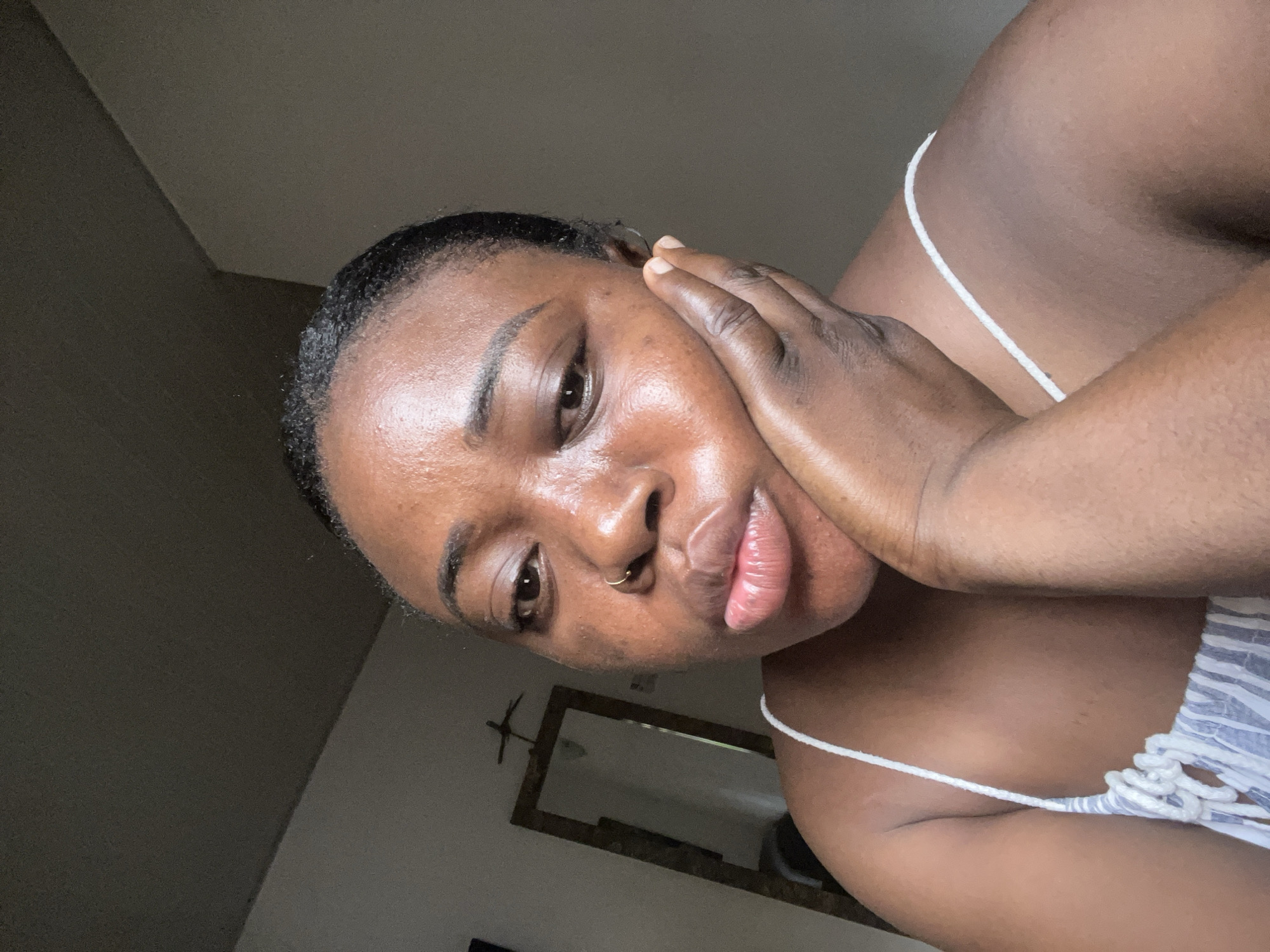 A medium-close-up shot, taken in the city of Abuja, Nigeria, shows a dark-skinned woman in a striped camisole. She faces forward, with her head tilted downward and to the left. Her left hand is cupped against the left side of her face. She has dark eyes and hair slicked back from her forehead. A gold nose ring is visible. The background shows a wall with a framed mirror.
