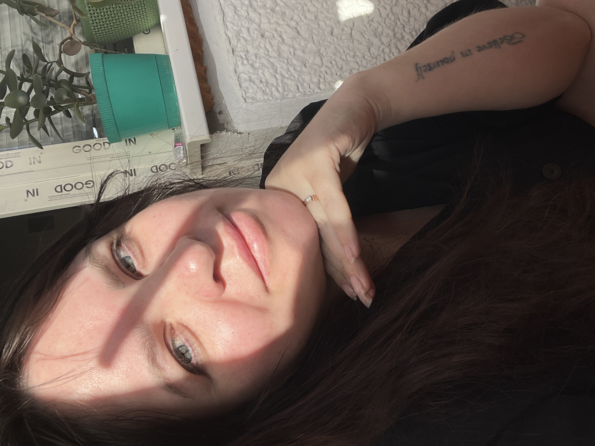 A woman with fair skin and dark brown hair is lying down, her head resting on her hand, which has a gold ring on the ring finger. She has a tattoo on her left arm that reads "Believe in yourself." The background includes a potted plant in a blue and green container, and a textured white surface. The word "GOOD" is visible in the background. The location is indicated as Noviy, Russia, but there are no specific elements in the image to verify this location forensically.