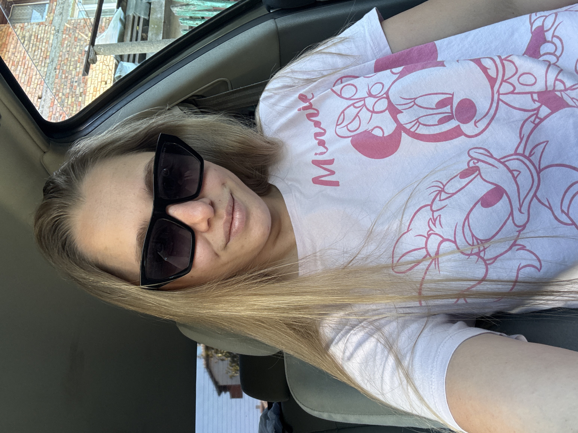 A blonde woman wearing sunglasses and a pink t-shirt with Minnie Mouse graphics is sitting in the driver's seat of a car. The car appears to be parked in a rural or semi-rural area. In the background, there is a brick building and a wooden fence. The sky is visible through the car window, suggesting it is daytime. The image was taken in Tsarevo, Bulgaria.
