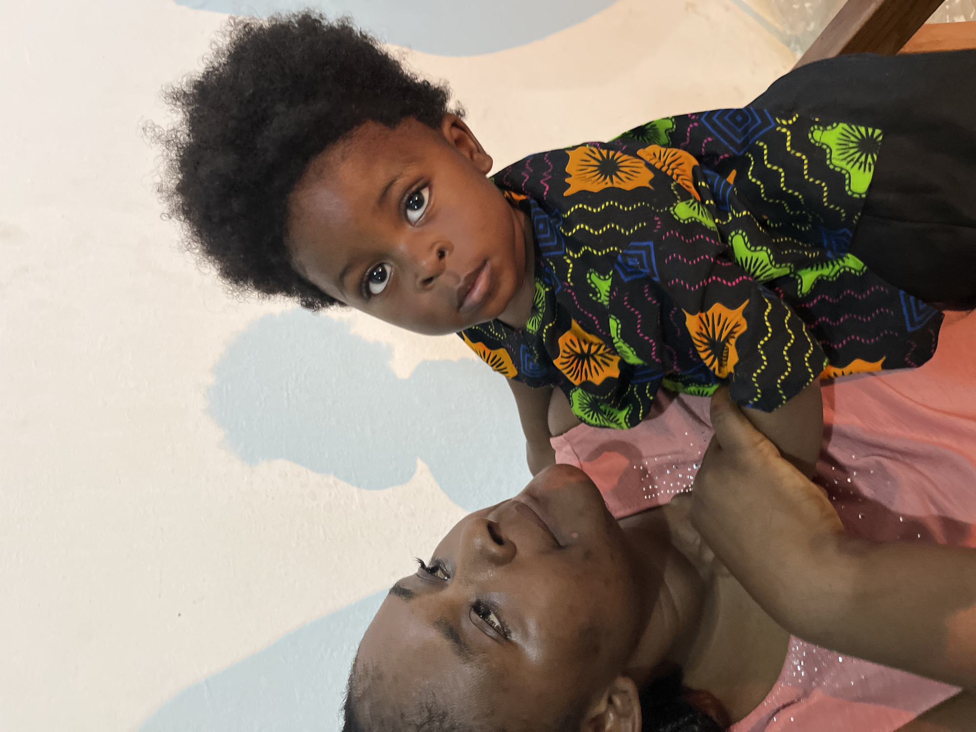A young child with dark, curly hair is held by an adult, who is wearing a pink shirt with rhinestones. The child is wearing a black shirt with a colorful pattern. The background is a plain wall, with shadows suggesting it is indoors. The lighting is soft, and the overall atmosphere is warm and intimate. The scene is likely set in a home in Gura Top, Nigeria.