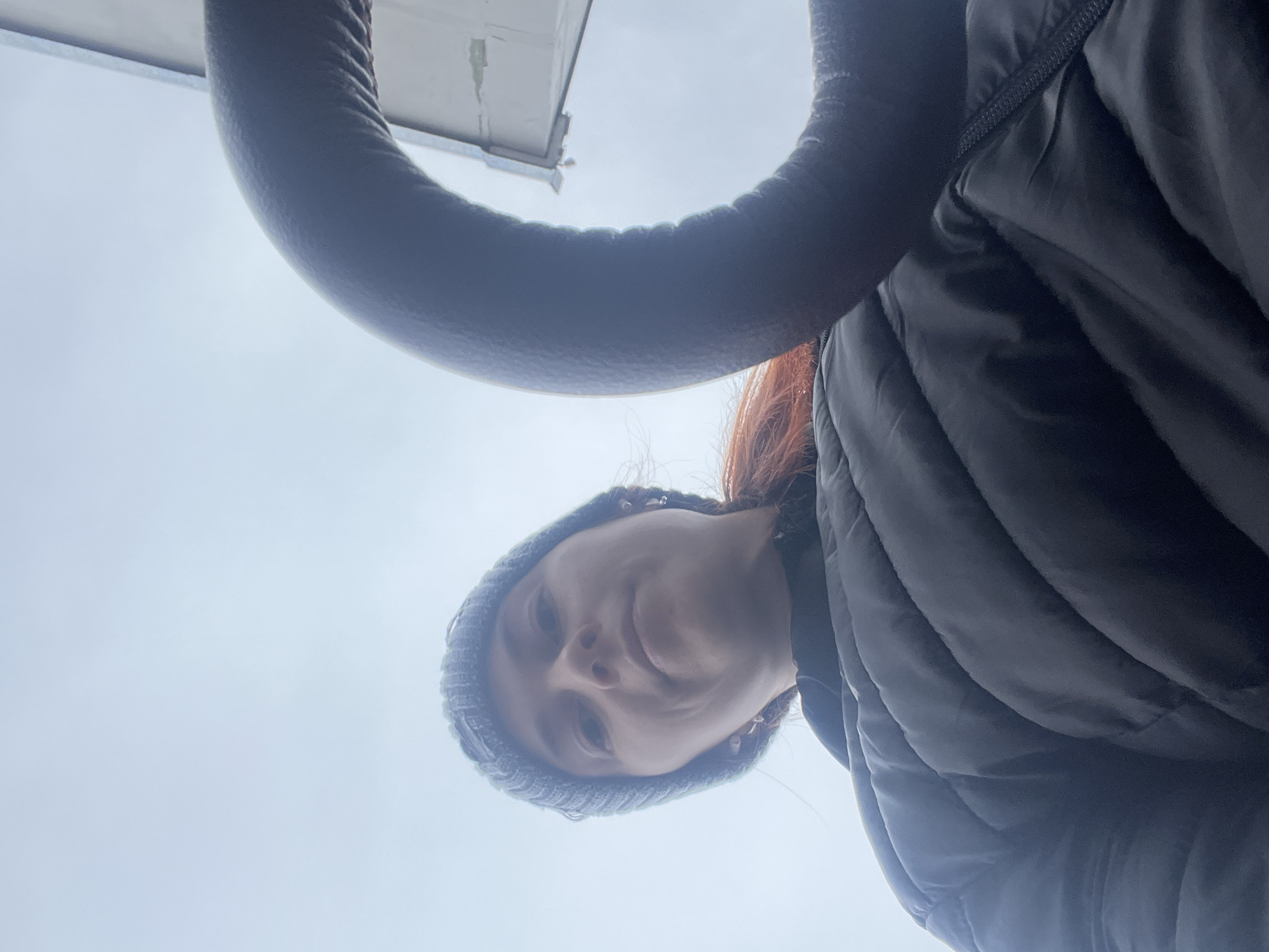A low-angle shot from below shows a person wearing a gray knit hat and a puffy black jacket. The sky is visible in the background, along with a gray curved object at the top left of the frame, possibly a handle or part of a vehicle. The person is looking down towards the camera. The location is possibly Chaikovsky, Russia.