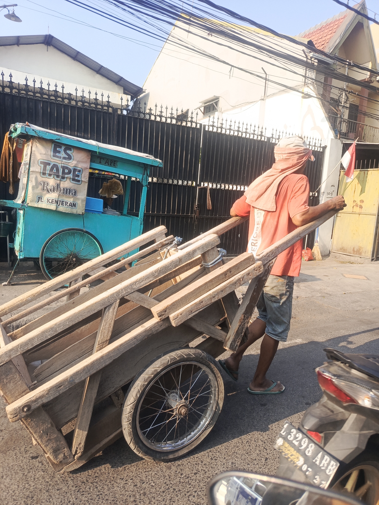 A man pushes a wooden cart down a street in Surabaya, Indonesia. The man is wearing a coral-colored t-shirt, denim shorts, and a head covering. He is pushing the cart by the handles, and it appears to be loaded with raw materials for construction. In the background, there is a teal-colored cart with a banner that reads "ES TAPE Rahma JL. KENTERAN". The scene is set in a street with a fence and buildings in the background, and a motorcycle is parked to the right of the frame. The sky is clear and blue, indicating a sunny day.
