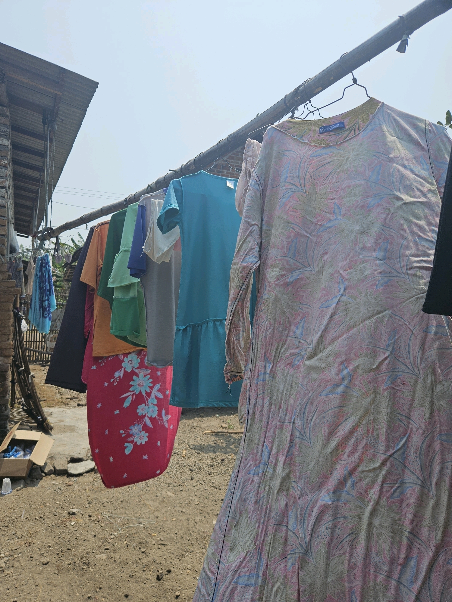 In Kampungbaru, Indonesia, laundry hangs on a clothesline outdoors. The clothesline is suspended between two poles, one made of bamboo and the other appearing to be a metal pipe. Several garments are visible, including a long-sleeved dress with a floral pattern in shades of pink, green, and blue, a bright turquoise dress, a pink skirt with a floral print, a dark blue shirt, an orange shirt, a green shirt, and a light gray shirt. Some smaller items, possibly towels or scarves, hang to the left. In the background, a corrugated metal roof shelters a brick structure, and a simple bamboo fence encloses the area. The ground is covered with dirt and small stones, and a cardboard box sits on the ground near the clothesline. The scene is illuminated by bright daylight, suggesting a sunny day.