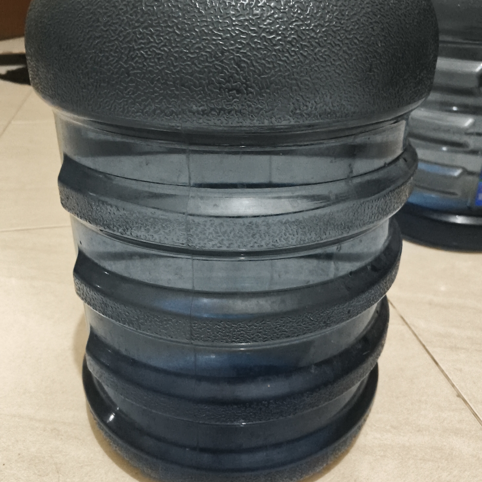 A close-up shot shows the textured top and ribbed body of a large, dark gray plastic water jug. The jug is resting on a tiled floor. Another similar water jug is partially visible in the background, to the right.