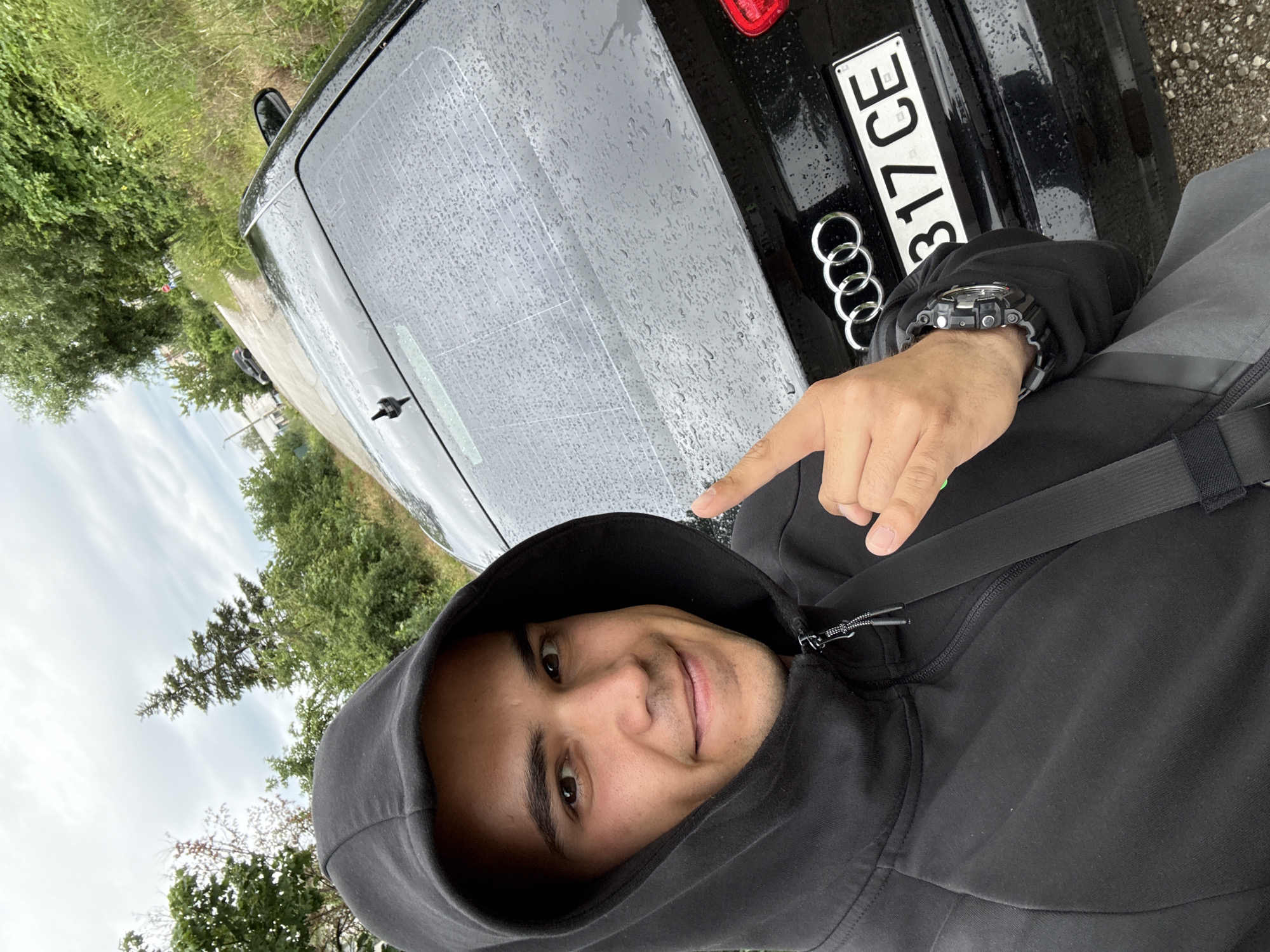 A close-up, wide-angle selfie shows a young man with dark hair and brown eyes wearing a black hooded sweatshirt. He is smiling and has his left hand raised, with his thumb and index finger extended in a gesture. The man is positioned in front of a black Audi car. The car's rear windshield is visible and appears to be wet, with raindrops evident. The license plate of the car reads "8317 CE". The background consists of trees and vegetation lining a paved road that curves away. The sky is overcast. The image is taken outdoors in what appears to be a rural or suburban area.