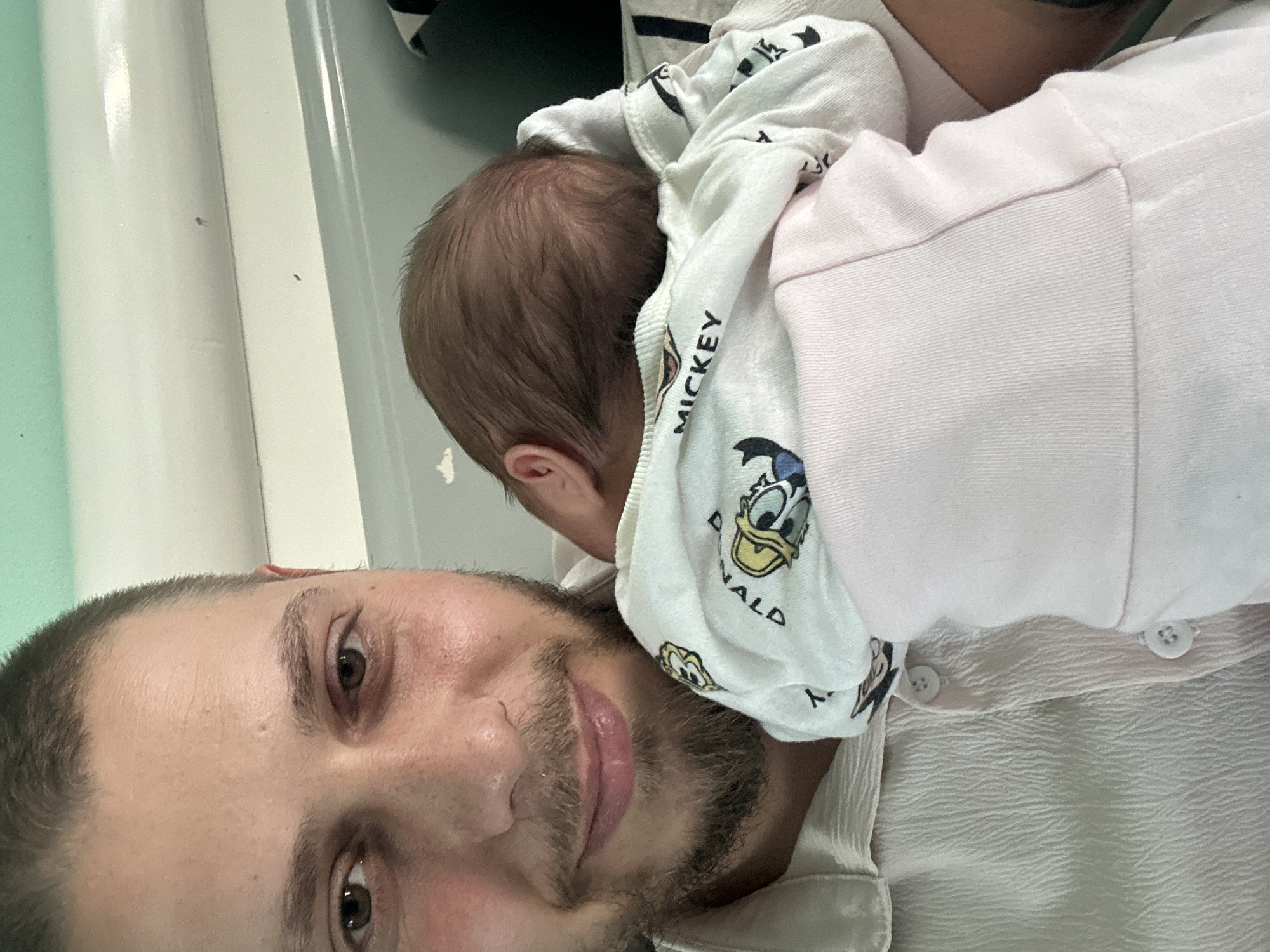 The image depicts a man holding a baby. The man has short brown hair and a beard, and he is wearing a light-colored button-down shirt. He appears to be smiling slightly. The baby is nestled against his chest, with their head turned away from the camera. The baby is wearing a pink shirt and a white blanket with cartoon character prints on it. The background is blurred, but it appears to be an indoor setting with light-colored walls. The location is not specified.