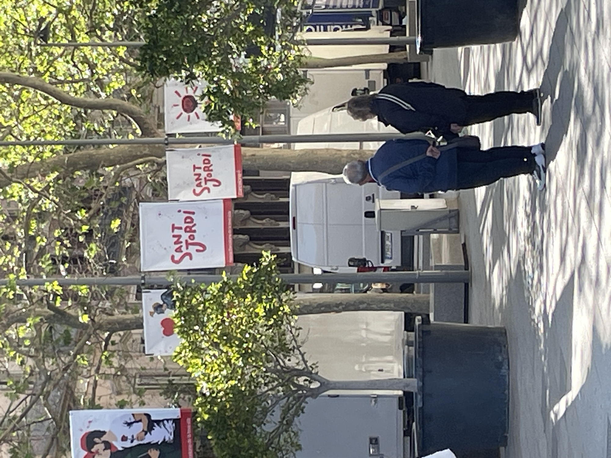 The image depicts a street scene in Barcelona, Spain. Banners advertising "Sant Jordi" are visible, suggesting the photograph was taken around April 23rd. Two people are walking on the sidewalk, and there are several white vans parked along the curb. Trees line the street, some in large planters. The scene is bathed in sunlight, creating sharp shadows on the sidewalk.