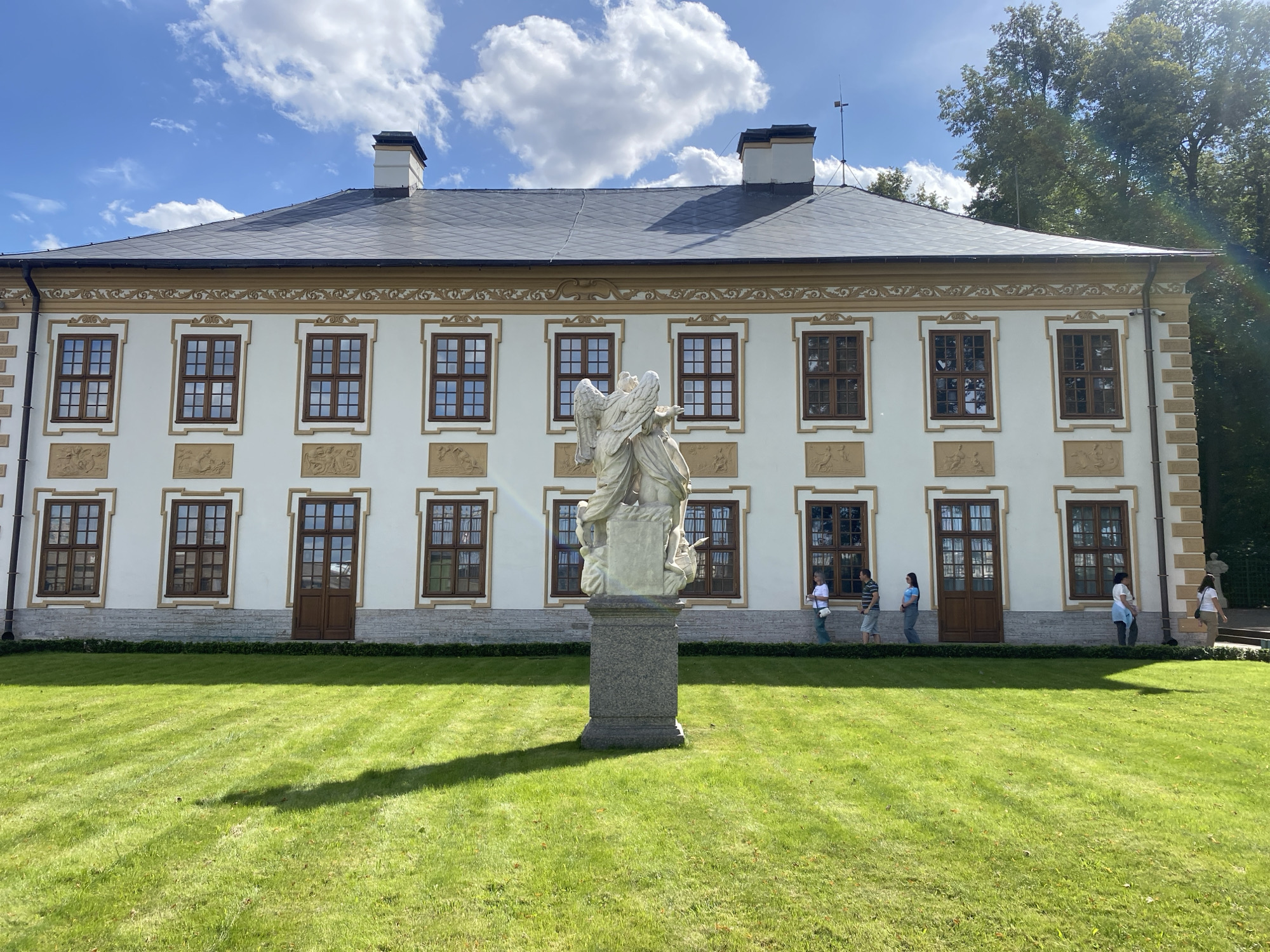 The image depicts a large, white building with a dark, pitched roof, flanked by trees on the right side. A white sculpture of an angel stands prominently in the foreground on a stone pedestal, casting a shadow on the well-maintained green lawn. The building features multiple windows with brown frames and decorative elements above and below them. Several people are visible walking in front of the building. The sky is blue with scattered white clouds. The architectural style and the presence of similar buildings and sculptures suggest this location is likely the Pavlovsk Palace complex in Saint Petersburg, Russia.