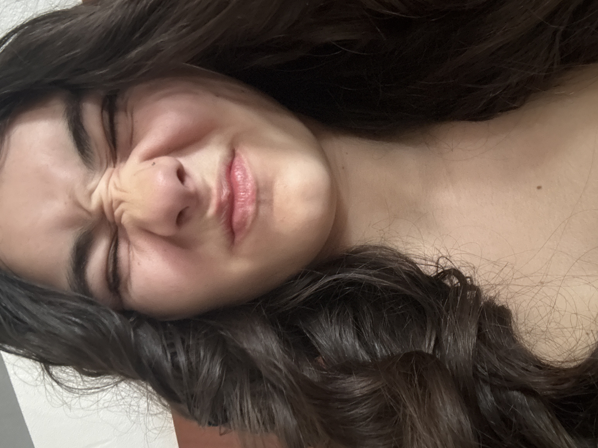 A close-up shot shows a woman's face contorted into a grimace. Her eyes are tightly shut, and her nose is wrinkled. Her mouth is slightly pursed. Dark brown hair frames her face. A mole is visible on her upper chest. The background is out of focus, but it appears to be a white surface. The image provides no evidence or indication that it was taken in Plovdiv, Bulgaria.