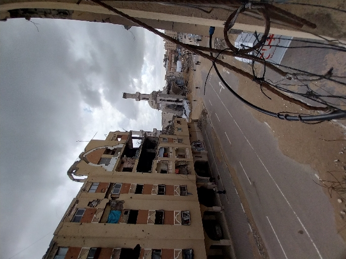 The image depicts a street scene in Khan Yunis, Palestinian Territory, characterized by extensive urban destruction under an overcast sky.
In the immediate foreground on the right, exposed rebar and entangled wires are visible, suggesting the vantage point is from a damaged structure. A two-lane asphalt road with double white lines and a directional arrow occupies the midground, its edges lined with sand and debris.
To the left of the road, a multi-story, light-brown residential building stands heavily damaged. Large sections of its facade are missing, exposing internal structural elements and rooms. Windows are shattered or entirely absent, and multiple circular impact marks, consistent with bullet holes, are visible on its exterior. A blue tarp hangs from a window opening on an upper floor.
Further down the street, a mosque with a tall minaret is partially destroyed, its dome and portions of its main structure visibly collapsed into rubble. Debris is scattered around its base and extends into the background, where more flattened or damaged buildings are discernible.
A few individuals are present on the scene. One person in dark attire walks along the sidewalk in front of the damaged apartment building. Several small figures are seen walking on the street and amidst the rubble near the mosque in the distance. On the right side of the street, a white banner with red text and a red emblem resembling a cross is partially visible. The sky is uniformly grey with heavy cloud cover.
