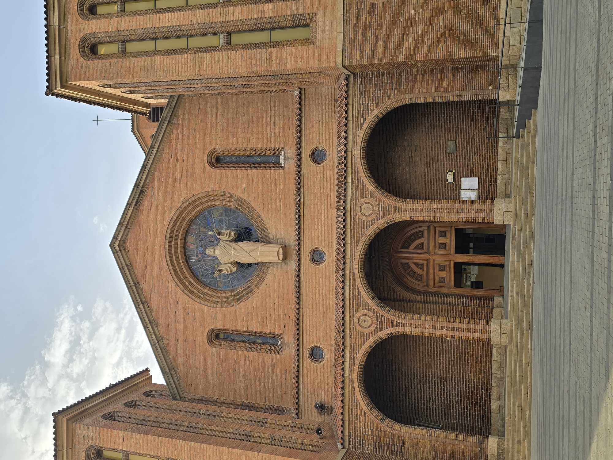 The image shows the facade of a brick church in Cornellà de Llobregat, Spain, under a cloudy sky. The church has a large, arched wooden double door at the entrance, flanked by two arched alcoves. Above the entrance, a statue of Jesus Christ is positioned in front of a circular stained-glass window. The facade is decorated with smaller arched windows and circular decorative elements. Two prominent brick towers flank the main facade, with tall, narrow windows. A thin cross is visible on a rooftop in the background. The foreground consists of a paved courtyard with cobblestone patterns. No people are visible in the image.