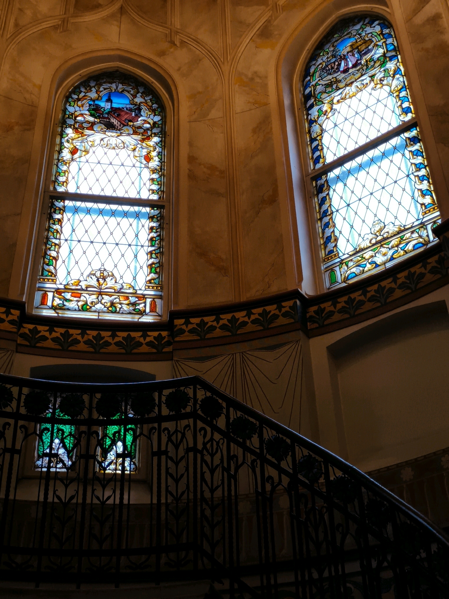 The image depicts a grand staircase with stained-glass windows in a building in Tallinn, Estonia. Two tall, arched stained-glass windows are visible on an upper level, illuminated by natural light. The window on the left features a scene of buildings and a blue sky, framed by intricate floral patterns. The window on the right also depicts a scene with figures and a blue sky, bordered by similar elaborate designs. A decorative frieze runs along the wall below these windows, adorned with dark leaf motifs.
In the foreground, a dark, ornate metal railing curves along the staircase, casting shadows. Below the railing, on a lower level, two smaller stained-glass windows with green and white patterns are visible, partially obscured by the railing. The walls of the building have a textured, yellowish-orange hue, with some subtle marbling. The overall impression is one of historical architecture and elegance, suggesting a location such as a museum, a historic hotel, or a public building with significant architectural heritage. The scene appears to be unoccupied by people.