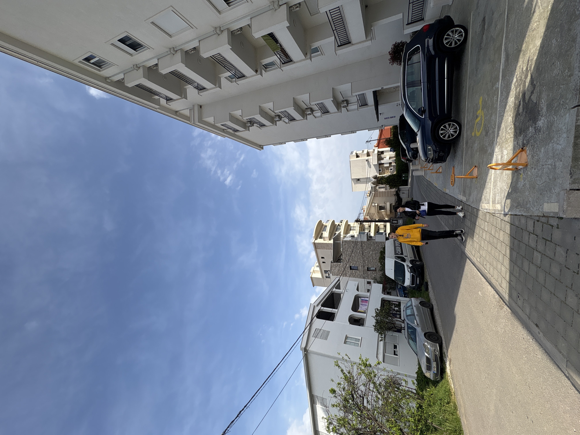 The photo depicts a street scene in Bar, Montenegro, lined with white buildings of varying heights and architectural styles. Two people, one wearing a yellow top, are walking along a paved road. Several parked cars are visible on both sides of the street, including a dark blue car in a designated parking space marked with a wheelchair symbol. Metal parking barriers are placed near the parked dark blue vehicle. A partly cloudy sky is visible above the buildings. Power lines run along the lower part of the frame. Vegetation is present along the roadside, including trees and grass.