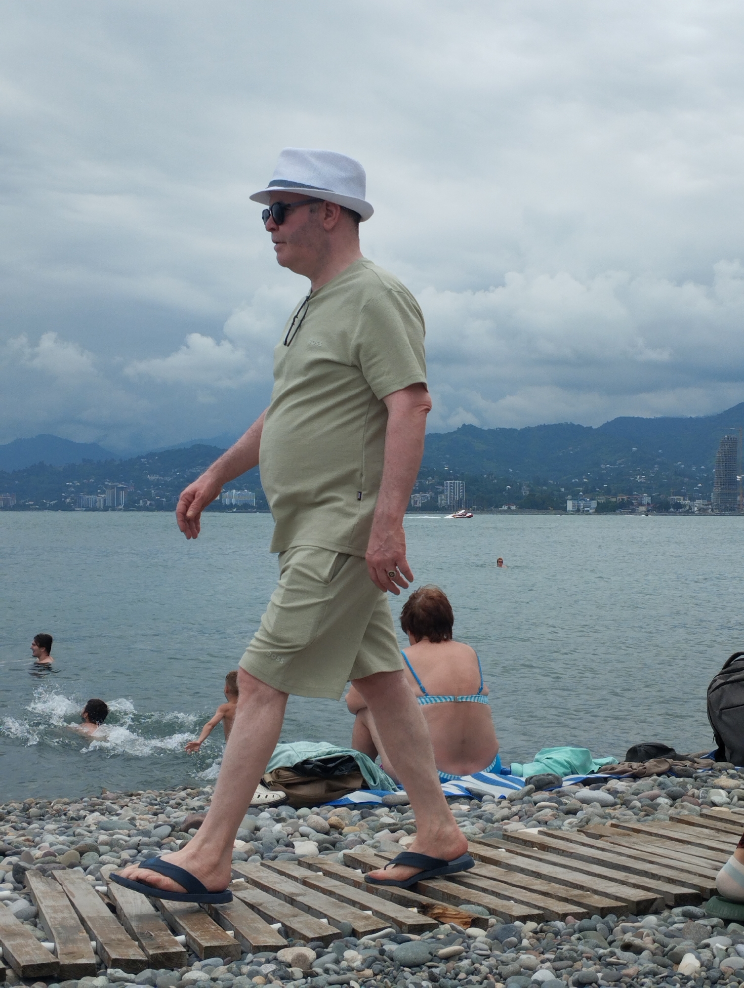 A man in a light-colored fedora hat and sunglasses walks along a wooden pathway on a pebble beach. He is wearing a light green t-shirt and matching shorts. He is walking towards the left of the frame. In the background, several people are in the water. A woman with brown hair is sitting on a towel on the beach, wearing a blue and white striped bikini. The sea is calm, and there are mountains in the distance with buildings and a tall skyscraper. The sky is overcast with dark clouds. There is a boat on the water. The overall atmosphere is relaxed and summery. The shorts have "BOSS" visible on them.