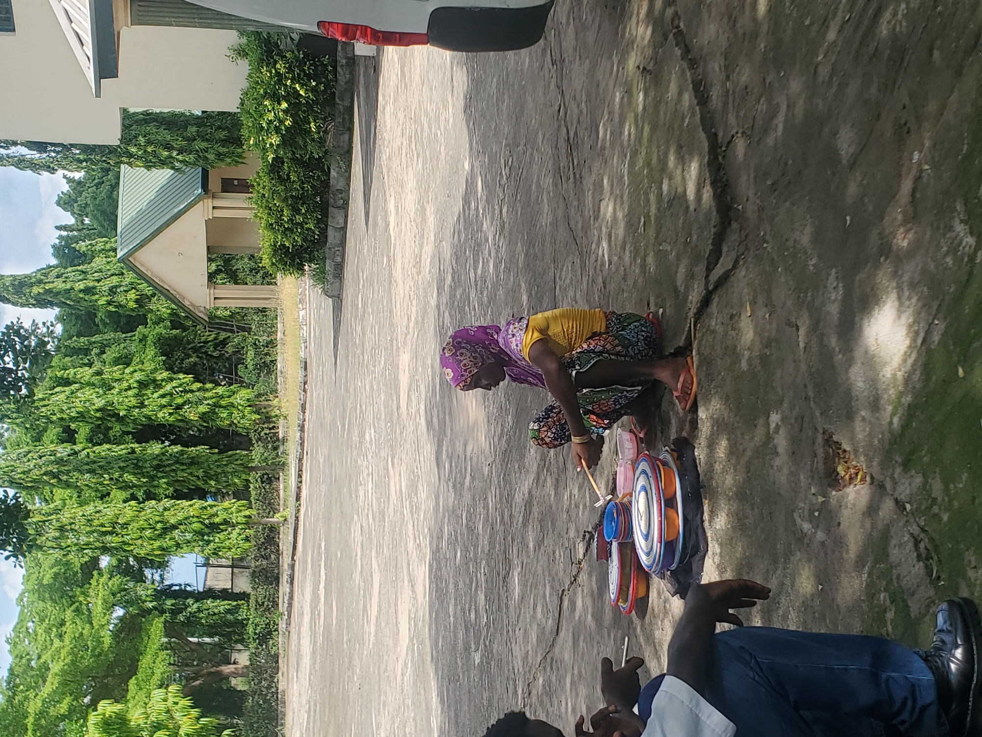 A woman wearing a purple headscarf and a colorful skirt crouches on a paved surface, interacting with a collection of brightly colored objects arranged on a dark cloth. She holds a thin stick, seemingly playing with or arranging these items. The objects appear to be circular and stacked, resembling toy drums or perhaps bowls. Next to her, another person is partially visible, wearing a blue shirt and dark pants.
The scene is outdoors, with dappled sunlight suggesting it's daytime, likely mid-morning or afternoon. The ground is a textured, cracked concrete or asphalt, with patches of green grass and shadows indicating trees and foliage nearby. In the background, a building with a green-tiled roof and cream-colored walls is visible, surrounded by lush, green trees. The overall atmosphere appears to be casual and everyday. There is no visible text in the image. The setting suggests a warm climate.