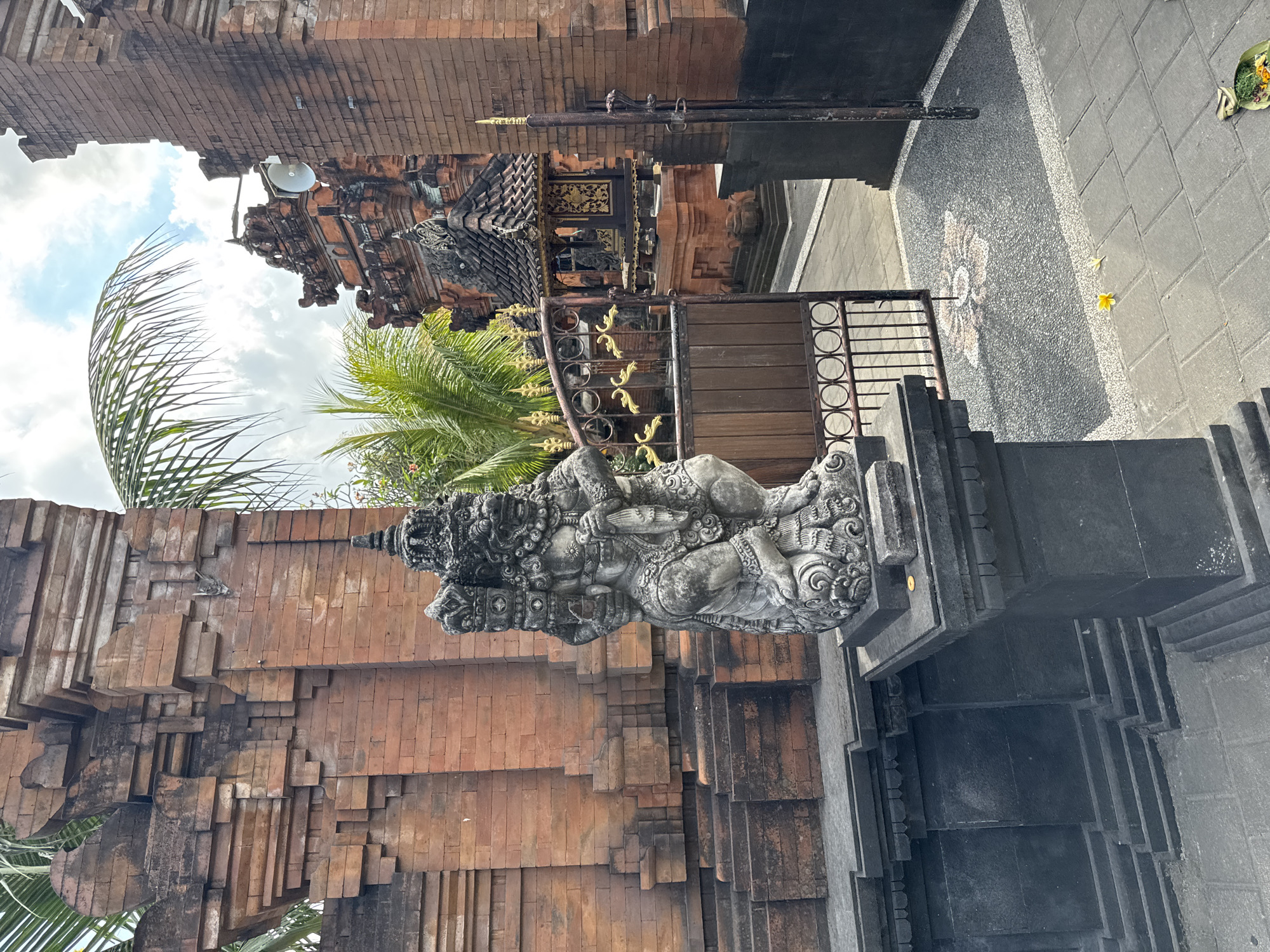 The image depicts an outdoor scene in Denpasar, Indonesia, featuring traditional Balinese architecture and religious elements. In the foreground, a weathered stone statue of a multi-armed deity stands on a dark, tiered pedestal. The statue is intricately carved, showing details of its facial features and elaborate attire. Behind the statue, a large brick structure with layered architectural details forms part of a temple complex.
Further into the scene, a decorative metal gate, adorned with gold accents and circular motifs, is partially open, revealing a pathway and another building within the complex. This inner building features a dark, tiled roof with ornate eaves and a facade decorated with intricate golden patterns. Palm fronds from lush green trees are visible, casting shadows and framing the sky, which is partly cloudy with a bright blue expanse. The ground is paved with stone tiles and sections of dark gravel. Two yellow flowers, likely offerings, lie on the paving stones, and a small, leaf-wrapped offering lies in the lower right corner. The overall impression is one of a serene, culturally rich, and spiritual environment.