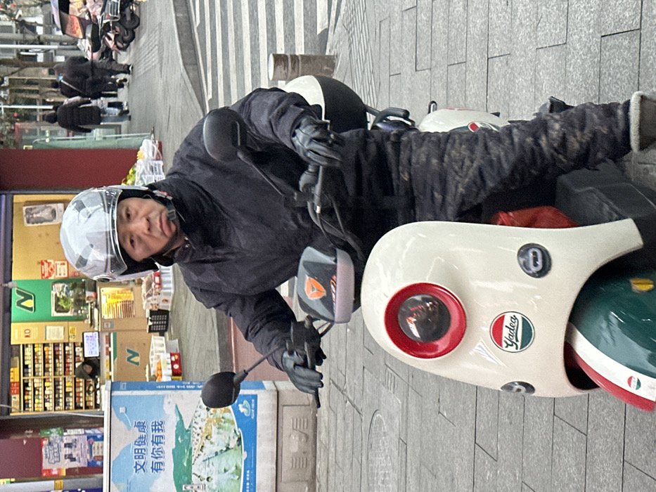 A middle-aged Asian male is depicted seated on an off-white Yadea electric scooter on a paved city street in Jiangjin, China. The individual wears a grey full-face helmet with a clear visor, a dark quilted jacket, matching dark quilted trousers, and black gloves, with his hands gripping the scooter's handlebars. His expression is neutral, and he looks directly forward. The scooter features a prominent red-rimmed circular headlight and a front fender with green, white, and red stripes; a "Yadea" logo is visible on the scooter's front panel.
In the background to the left, a retail storefront with a wide entrance is visible, displaying numerous products on shelves and featuring multiple green "N" logos. A person is dimly visible inside the store. Adjacent to the store, a light blue sign in Chinese reads "文明健康 ་ 你有我" (Civilized and healthy, with you and me) above a landscape image. The background to the right shows a grey tiled sidewalk with several blurred pedestrians walking in the distance, alongside parked scooters and a dark asphalt street with faint crosswalk markings. The scene is illuminated by natural light, consistent with an overcast day in an urban environment.