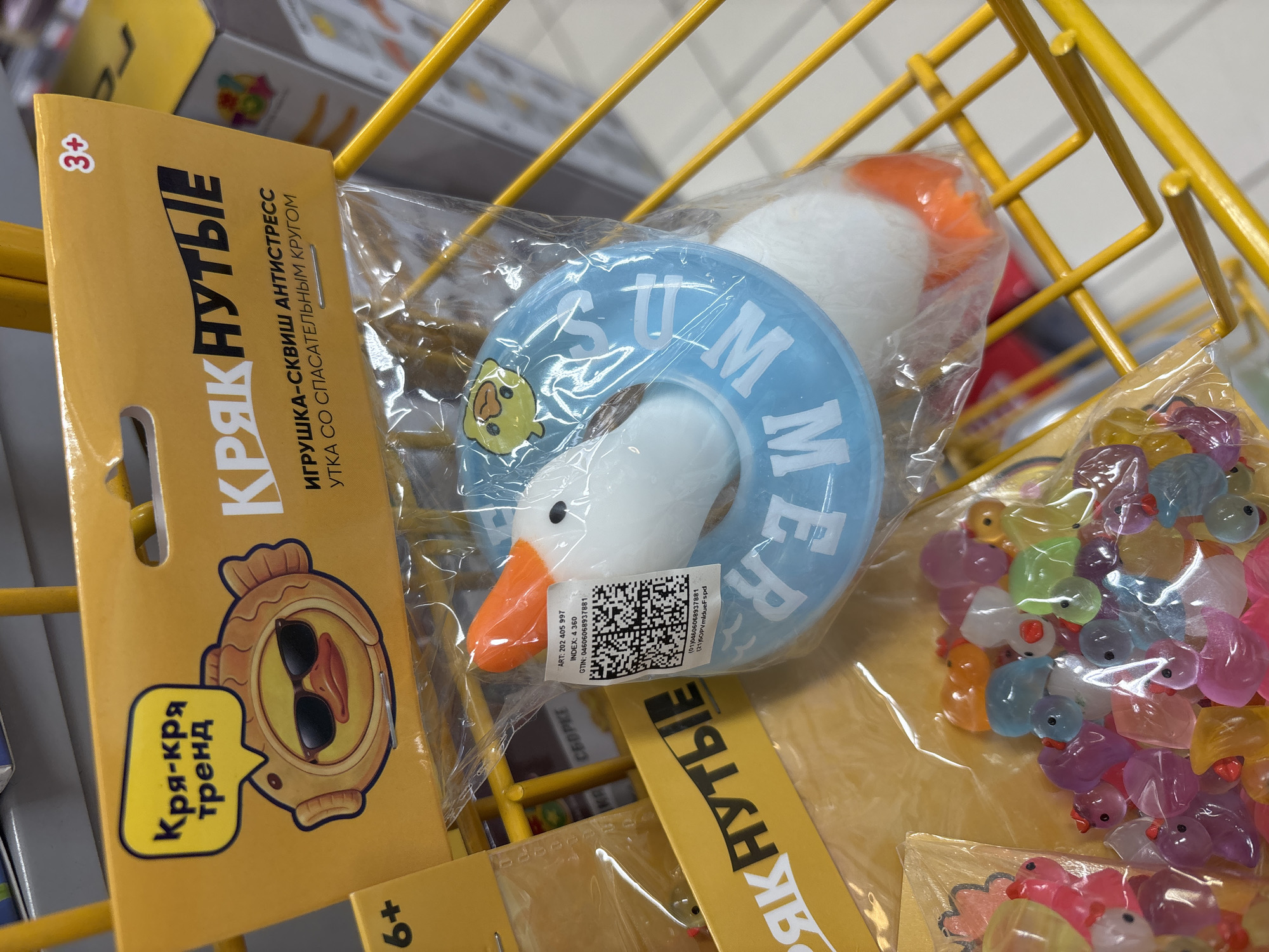 A close-up shot captures a display of novelty toys, likely for children, positioned within a yellow metal wire shopping cart. The primary focus is on a large, white toy duck with an orange beak, enclosed in clear plastic packaging. This duck is adorned with a blue inflatable ring around its neck, featuring white lettering that appears to spell "JUM" or "UM". The packaging is attached to a bright yellow header card displaying text in Russian. One prominent phrase on the card reads "Кря-кря тренд" (Quack-quack trend), accompanied by a cartoon image of a duck wearing sunglasses and a fish-shaped costume. Another section of the header card features larger, stylized Russian text that appears to be the product name "КРЯКНУТЫЕ" (Quacky). Below this, there is text indicating "ИГРУШКА-СКВИШ АНТИСТРЕСС" (Squeeze toy anti-stress) and "УТКА СО СПАСАТЕЛЬНЫМ КРУГОМ" (Duck with a life preserver). Age recommendations, "3+" and "6+", are also visible on the header card.
To the left of the main duck toy, another smaller, clear plastic bag holds an assortment of tiny, colorful, translucent duck-shaped toys in various shades of pink, purple, blue, green, and orange. Some of these smaller ducks have red beaks.
The shopping cart, with its bright yellow wire construction, is visible in the foreground and extends into the background. The background is blurred but suggests a retail environment, possibly a supermarket or toy store, with other product packaging and shelving faintly discernible. The lighting is bright and appears to be overhead artificial light.
The image was taken at the "Auchan" hypermarket located at 85B Kommuny Street, Saint Petersburg, Russia.