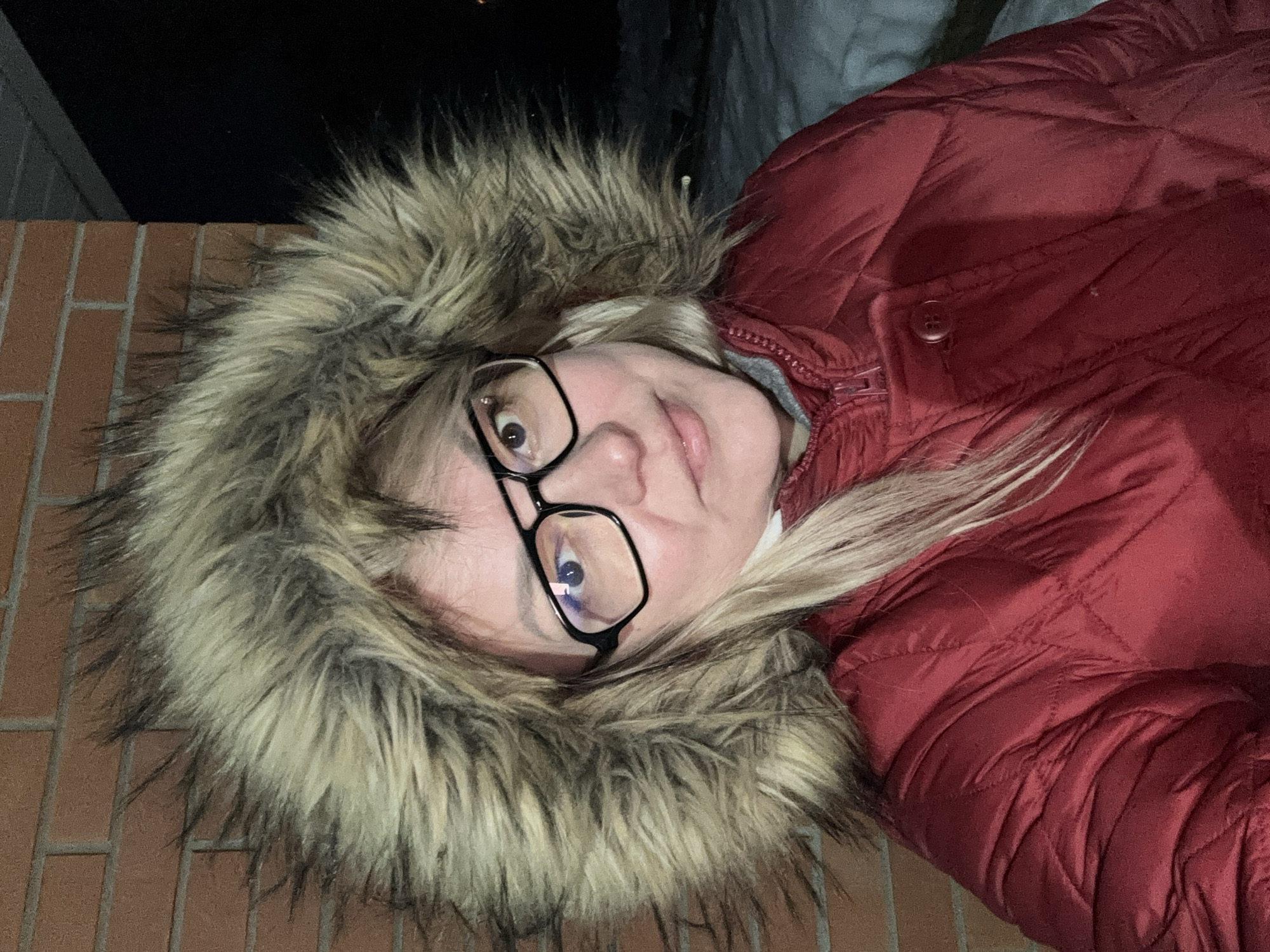 A close-up, chest-up portrait of an adult female in Русский Вожой, Russia, captured outdoors at night. The subject has fair skin, light blonde hair partially visible beneath a prominent, multi-toned light brown and dark brown/black faux fur hood, and wears dark-rimmed eyeglasses. Her gaze is directed towards the camera with a neutral expression. She is dressed in a red, quilted, insulated parka, which is partially zippered and features a buttoned storm flap. To the subject's left, a wall constructed of reddish-orange brickwork is visible. To her right, a layer of snow covers the ground, and the background is indistinctly dark, consistent with nighttime conditions. Artificial illumination highlights the subject's face and upper body.