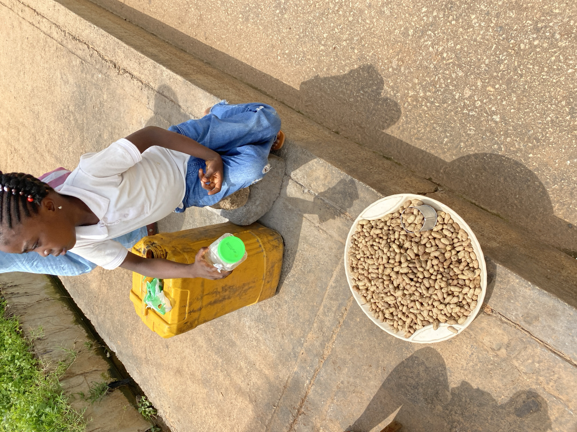 A young girl with braided hair and red beads, wearing a white polo shirt and ripped blue jeans, is crouching beside a large yellow plastic container. She is holding a clear plastic bottle with a green lid. In front of her, on the ground, is a white bowl filled with peanuts, and a small metal scoop is resting in the bowl. The girl appears to be engaged in some activity related to the peanuts or the container. The scene is set on a concrete surface, possibly a sidewalk or roadside, with a drainage ditch and green vegetation visible in the background. Sunlight casts distinct shadows, indicating an outdoor environment. The location is identified as Bukuru, Nigeria.