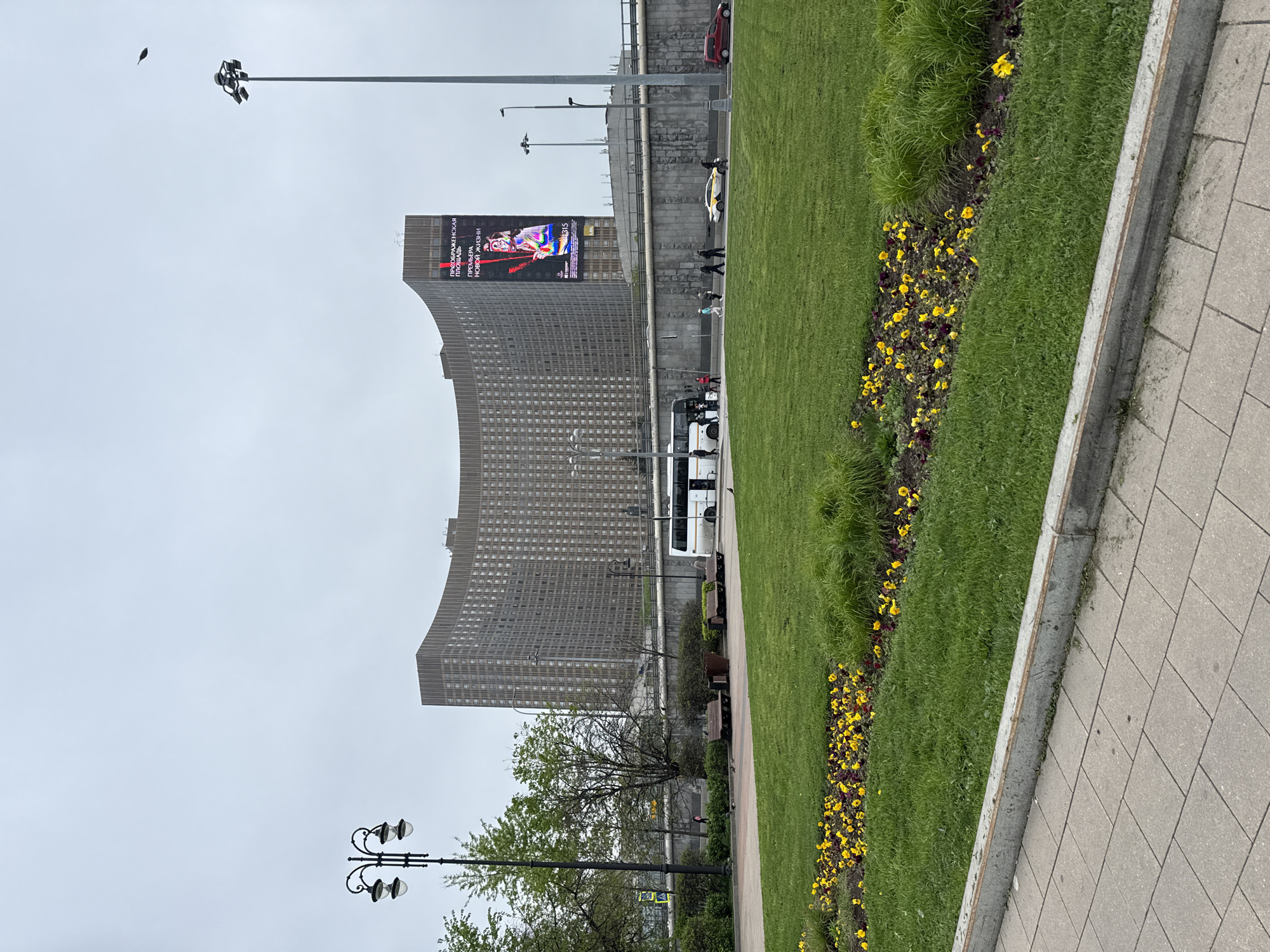 The image shows a cityscape in Moscow, Russia, dominated by the Hotel Leningradskaya (Hilton Moscow Leningradskaya), a Stalinist skyscraper. A large advertisement is displayed on the upper section of the building. The ground level features a grassy area with flowerbeds containing yellow and purple flowers. A pedestrian walkway runs along the edge of the grass. A lamppost is visible in the foreground, and other streetlights are placed throughout the scene. A bus is parked near the hotel. Several people are walking on the sidewalk adjacent to the road. The sky is overcast.
