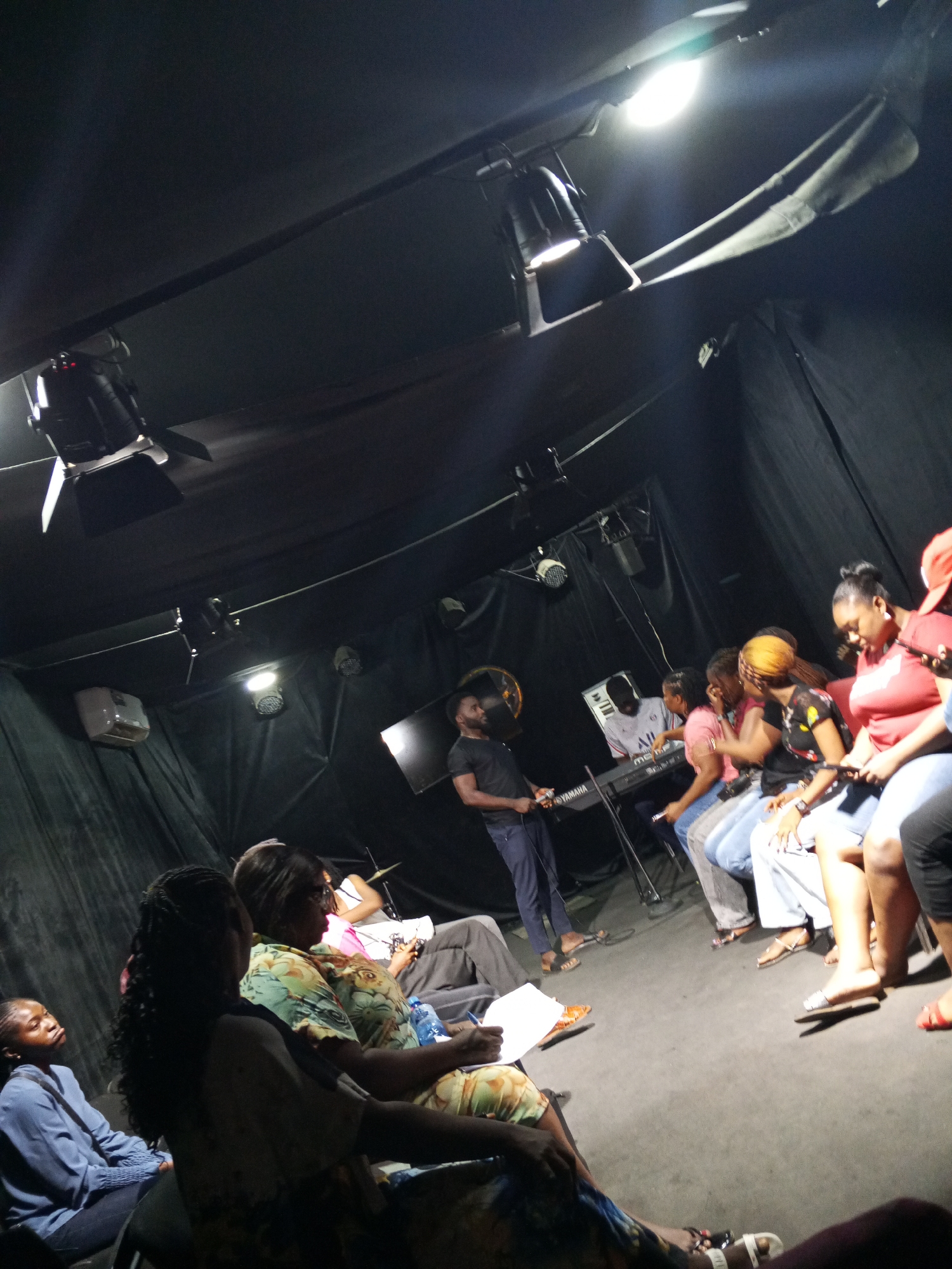 A dimly lit room, possibly a small theater or studio, is the setting for an event. A group of people sits in chairs, arranged in a semi-circle, suggesting an audience or participants in a workshop or performance. At least 14 individuals are visible. A man stands near a Yamaha keyboard, possibly addressing the group or preparing to play. Overhead, studio lighting fixtures are noticeable, indicating a controlled environment. The backdrop appears to be dark fabric, adding to the intimate atmosphere. The scene suggests a gathering focused on music, performance, or instruction in an indoor location. While not definitively shown, based on context, the location is likely in the city of Abuja, Nigeria.
