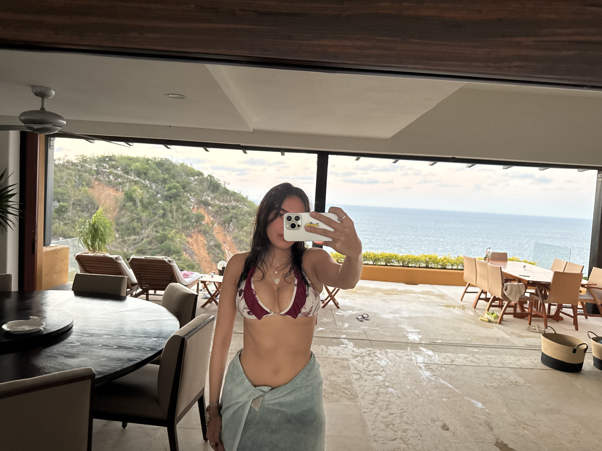 A young woman takes a mirror selfie in a luxurious, open-concept tropical setting. She stands in the foreground, facing away from the camera's perspective, holding a white smartphone to capture her reflection. Her face is obscured by the phone. She has long, dark hair, wears a red and white patterned bikini top, a silver necklace, and a light blue towel wrapped around her waist. A yellow sticker is visible on the back of her phone.
The setting transitions from an indoor dining area to an expansive outdoor patio, offering a breathtaking view of the ocean and a lush, green, mountainous coastline. In the immediate foreground to the left, a large, dark wooden round dining table with several upholstered chairs is visible, with a small white dish on it. Overhead, a modern ceiling fan hangs.
The outdoor patio, which appears wet in places, features several lounge chairs on a raised section to the left, and a rectangular dining table with chairs further out to the right. Two woven baskets with black bases are on the patio floor, along with a pair of flip-flops. Beyond the patio, the calm, blue ocean stretches to the horizon under a bright sky with scattered clouds. The background shows verdant hillsides characteristic of a coastal, tropical region. The scene suggests a relaxed vacation environment, likely during late morning or early afternoon.