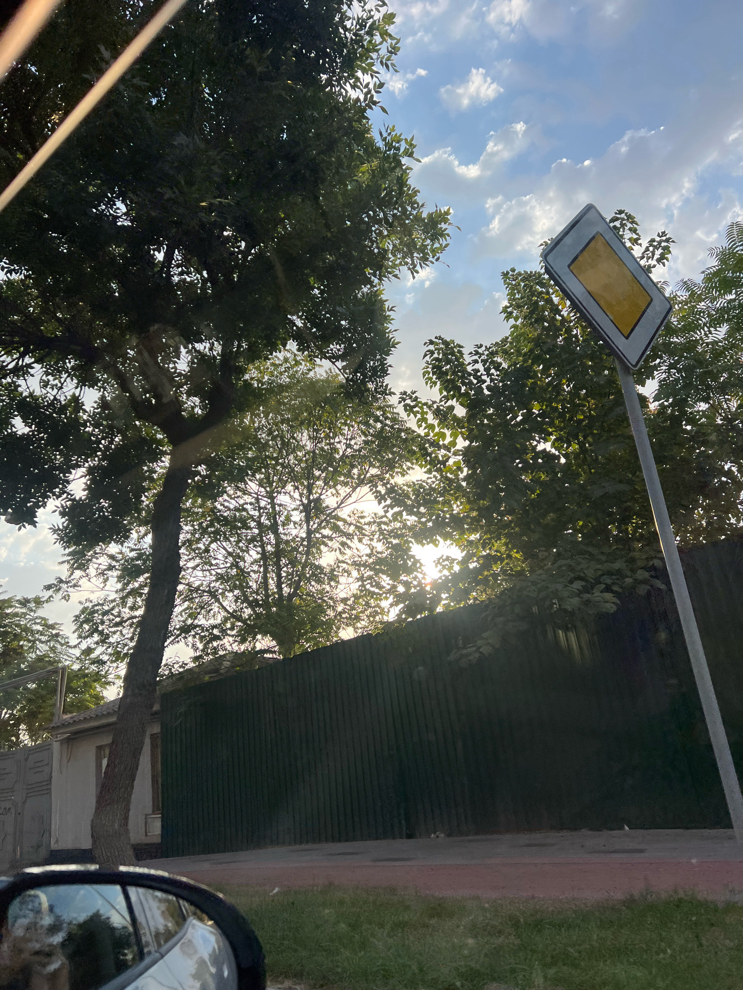 The image shows a street scene with trees, a fence, a building, and a road. The sun is shining through the trees, creating a dappled light effect. In the foreground, a car's side mirror is visible, reflecting the trees and sky. A road sign with a yellow rectangle inside a grey rhombus is mounted on a pole to the right of the image. A green corrugated metal fence runs along the side of the road, behind which a small building with a white facade and a grey gate are partially visible. The sky is blue with white clouds. The scene appears to be a residential area on a sunny day.