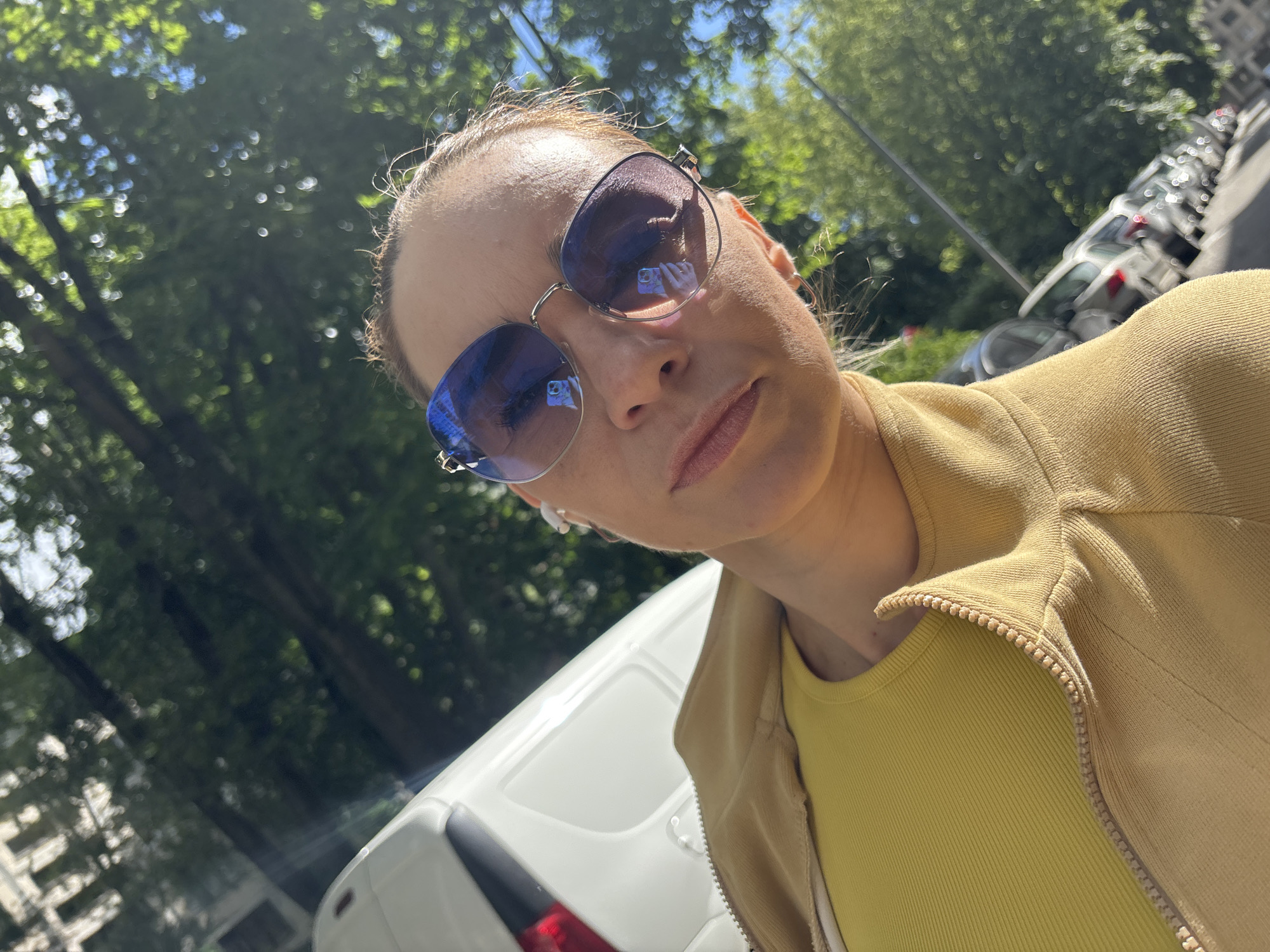 The image is a selfie taken outdoors on a sunny day. The subject is a woman wearing sunglasses and a yellow top under a tan zip-up jacket. Her hair is pulled back, and she is wearing earrings. The background shows dappled sunlight filtering through green trees, and a row of parked cars. In the foreground, the top of a white van is visible. The woman's expression is neutral, and the lighting suggests it is daytime. The context of Saint Petersburg, Russia, indicates an urban environment with trees lining a street where cars are parked.