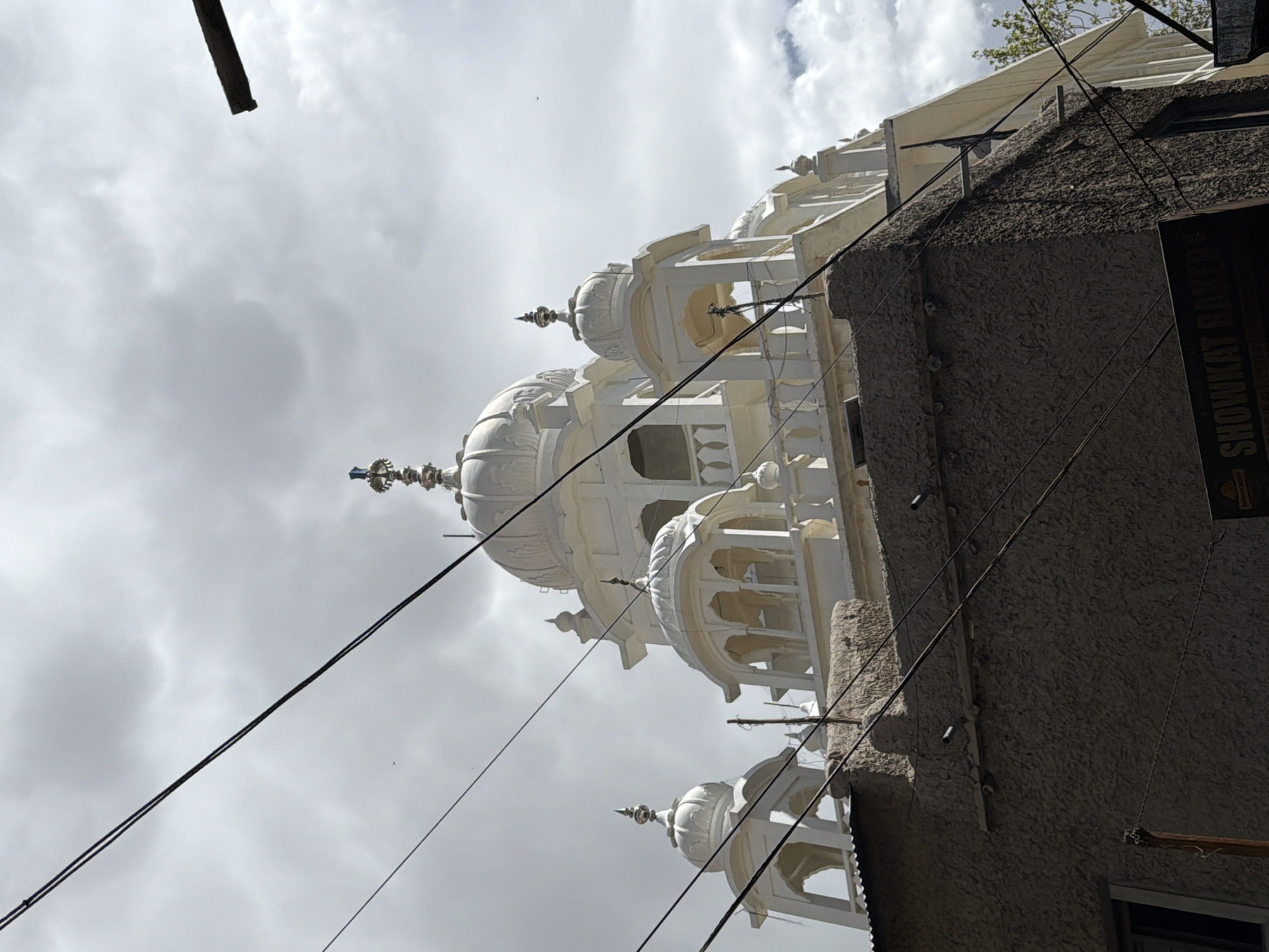 The image captures a white domed building with multiple smaller domes, likely a religious structure, against a cloudy sky. The building features intricate architectural details, including arches and spires. In the foreground, a darker, textured building partially obscures the view of the white structure. Several power lines crisscross the frame, casting shadows and adding a sense of urban complexity. The location is identified as the city of Leh, India, specifically a Sikh Gurudwara. A sign for "SHOWKAT BAKERY" is visible on the lower part of the frame, suggesting commercial activity nearby. The overall impression is one of a juxtaposition of spiritual architecture with everyday urban elements and utilities.