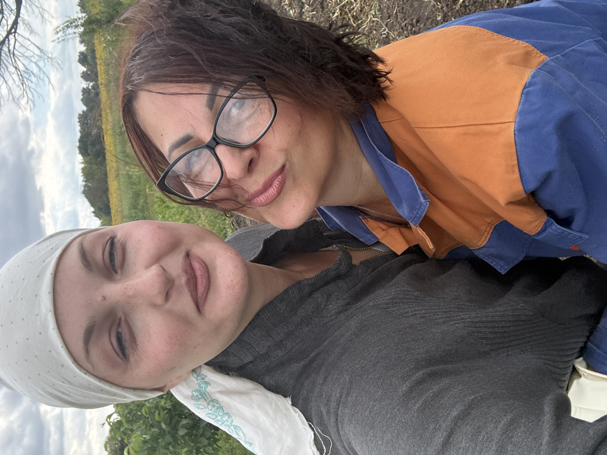 Two women are posing for a selfie outdoors. The woman on the left wears a white headscarf with a blue polka dot pattern and a grey cardigan. She has freckles on her face. The woman on the right wears glasses and an orange and blue jacket. They are standing in a rural area with dirt ground and vegetation in the background. The sky is cloudy. The location is Fominka, Russia.