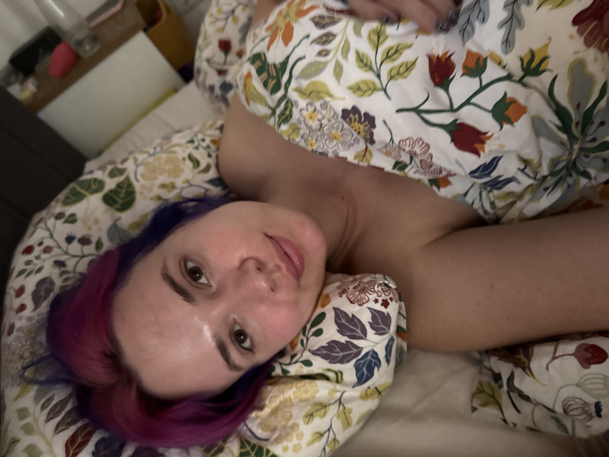 An indoor shot captures a woman lying in bed, looking directly at the camera from a slightly upward angle. Her head rests on a pillow, and she is partially covered by a duvet, both featuring a distinct floral pattern of leaves and flowers in shades of red, orange, yellow, green, blue, and purple on a light background. The woman has striking bi-colored hair, transitioning from purple near her scalp to vibrant pink or magenta towards the ends. Her face is bright and clear, with open eyes and a subtle, relaxed expression that hints at a gentle smile. Her lips are slightly parted. One of her bare shoulders and upper arm are visible from beneath the covers, indicating a state of undress or comfortable sleepwear.
In the blurred background on the left, elements of a nightstand or bedside table are visible. These include a clear water bottle, a pink object (possibly a sponge or small item), a yellow container or basket, and a white rectangular object, which could be a book or electronic device. The lighting in the room is soft and ambient, suggesting either early morning, late evening, or simply subdued indoor illumination, contributing to a calm and intimate atmosphere. The woman appears at ease and comfortable in her surroundings, possibly having just woken up or preparing to rest. No text is visible in the image. The individual is situated in the context of the City of Zagreb, Croatia.