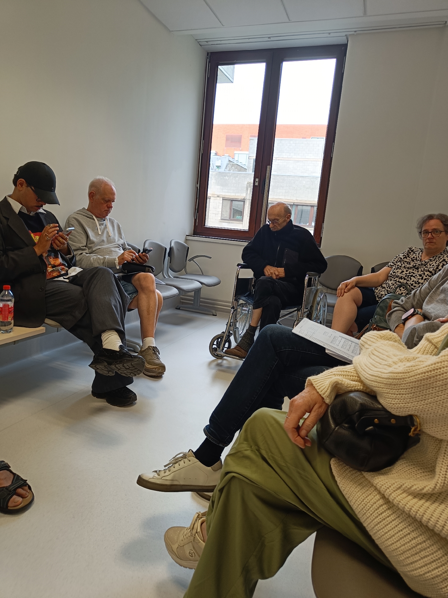 In a brightly lit indoor waiting area, possibly a medical clinic or administrative office, five individuals are seated, with one in a wheelchair. The environment is minimalist, with white walls and a large window overlooking an urban landscape with brick buildings.
On the left, a young man in a dark suit and baseball cap is looking at his phone. Next to him, an older man in a grey hoodie is also engrossed in his phone. Further along the bench, a man in a wheelchair wearing a dark jacket is looking down, and a woman with glasses in a patterned blouse is seated to his right, looking towards the viewer. In the foreground, a person wearing a cream-colored sweater and green pants is seated, with their handbag on their lap. The floor is a light-colored linoleum.