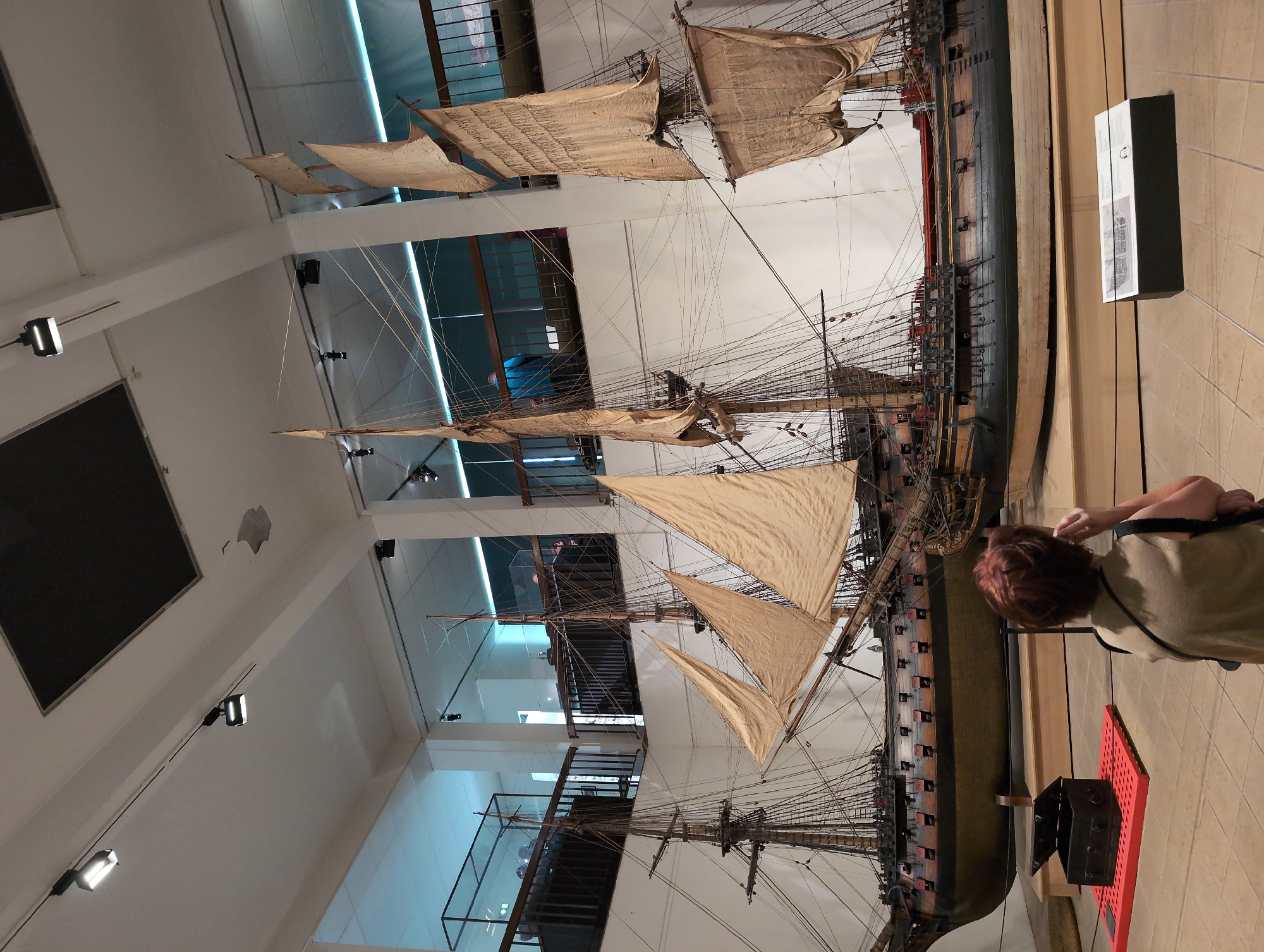 Here is a detailed description of the image:
The image depicts a large model ship inside a museum, likely in Toulon, France. The ship is a historic sailing vessel, meticulously detailed with masts, rigging, and sails. A visitor, partially visible on the right, observes the ship. The museum environment includes high ceilings, display cases, and architectural details. The lighting is bright, illuminating the ship and its surroundings. The ship model is positioned as a central focus, likely a significant exhibit. A display with a black box is on the floor, near the ship. The ship's location is the Marine Museum of Toulon.